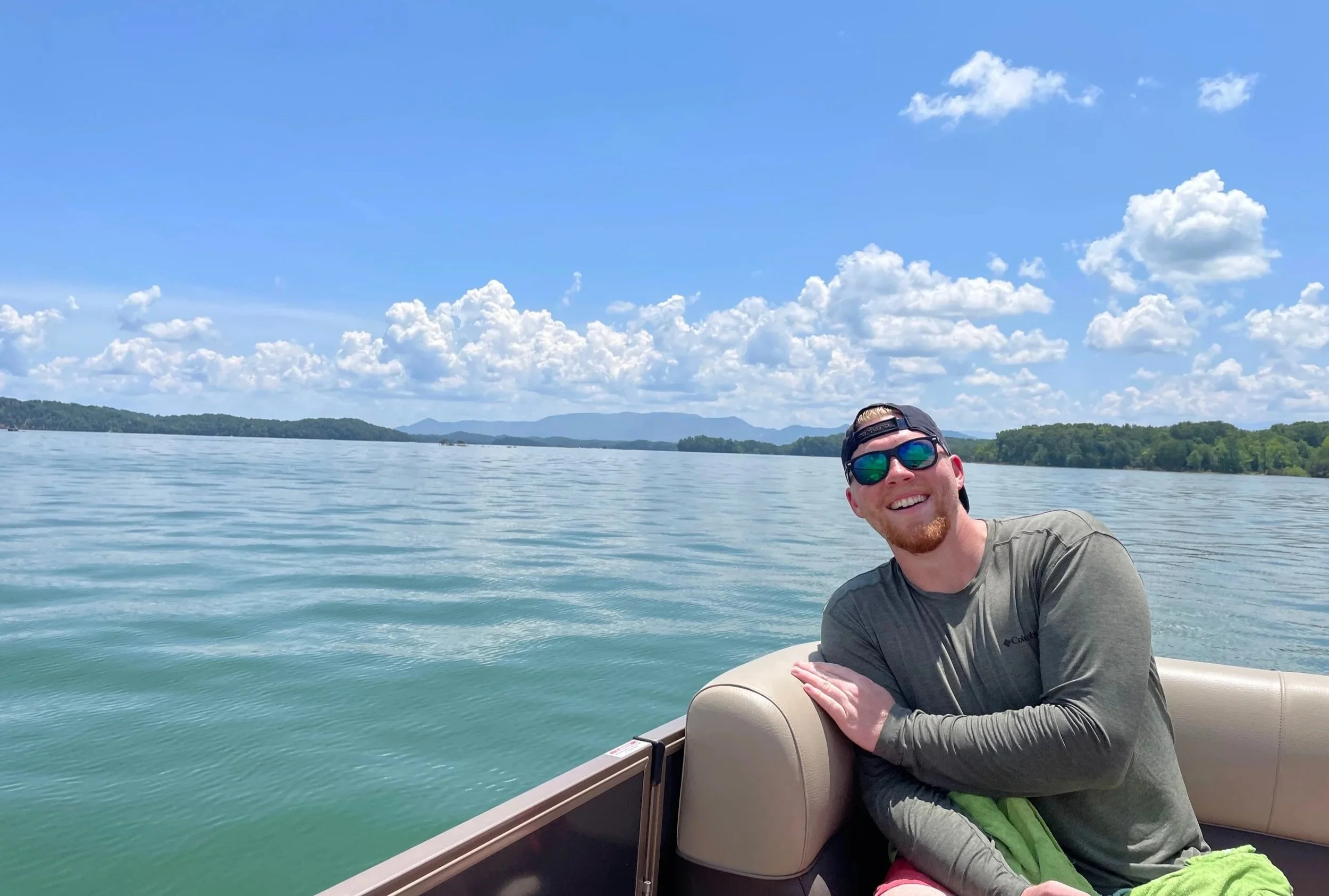 Smiling man on a boat during a sunny day with blue sky, white clouds, and distant mountains.