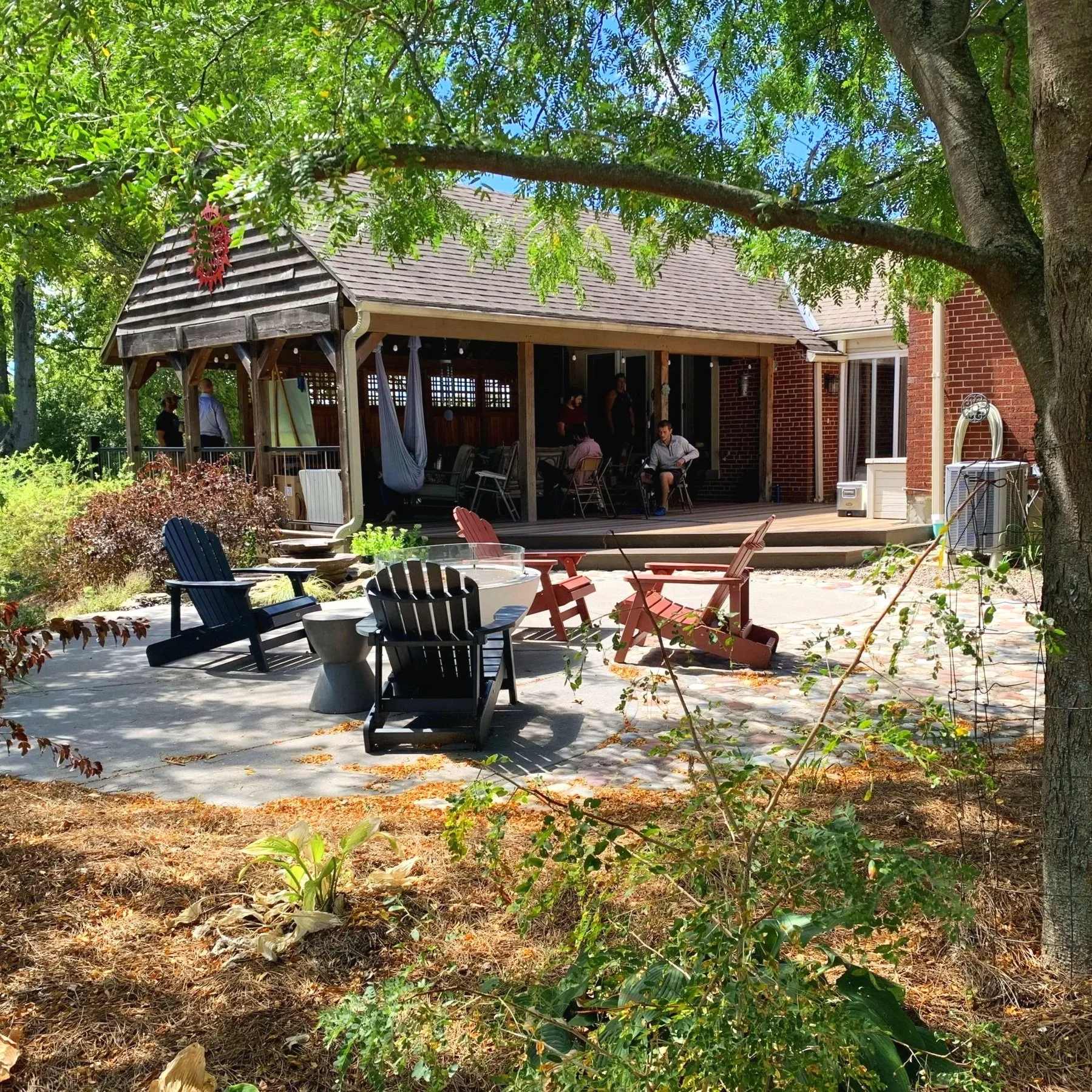 A backyard patio with chairs and trees, showing a house with a brick exterior and a porch with people sitting and standing.