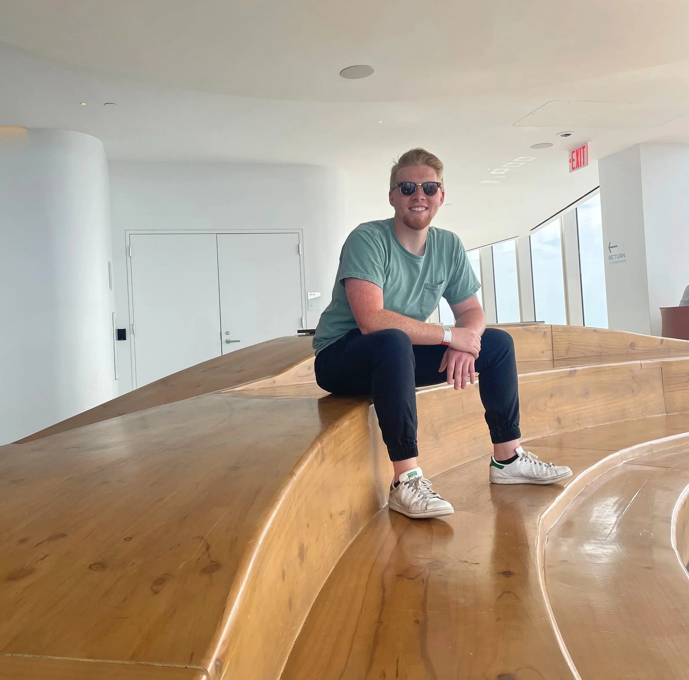 A young man with sunglasses, a green t-shirt, and black pants sitting on a curved wooden bench in a modern, well-lit interior space with large windows.