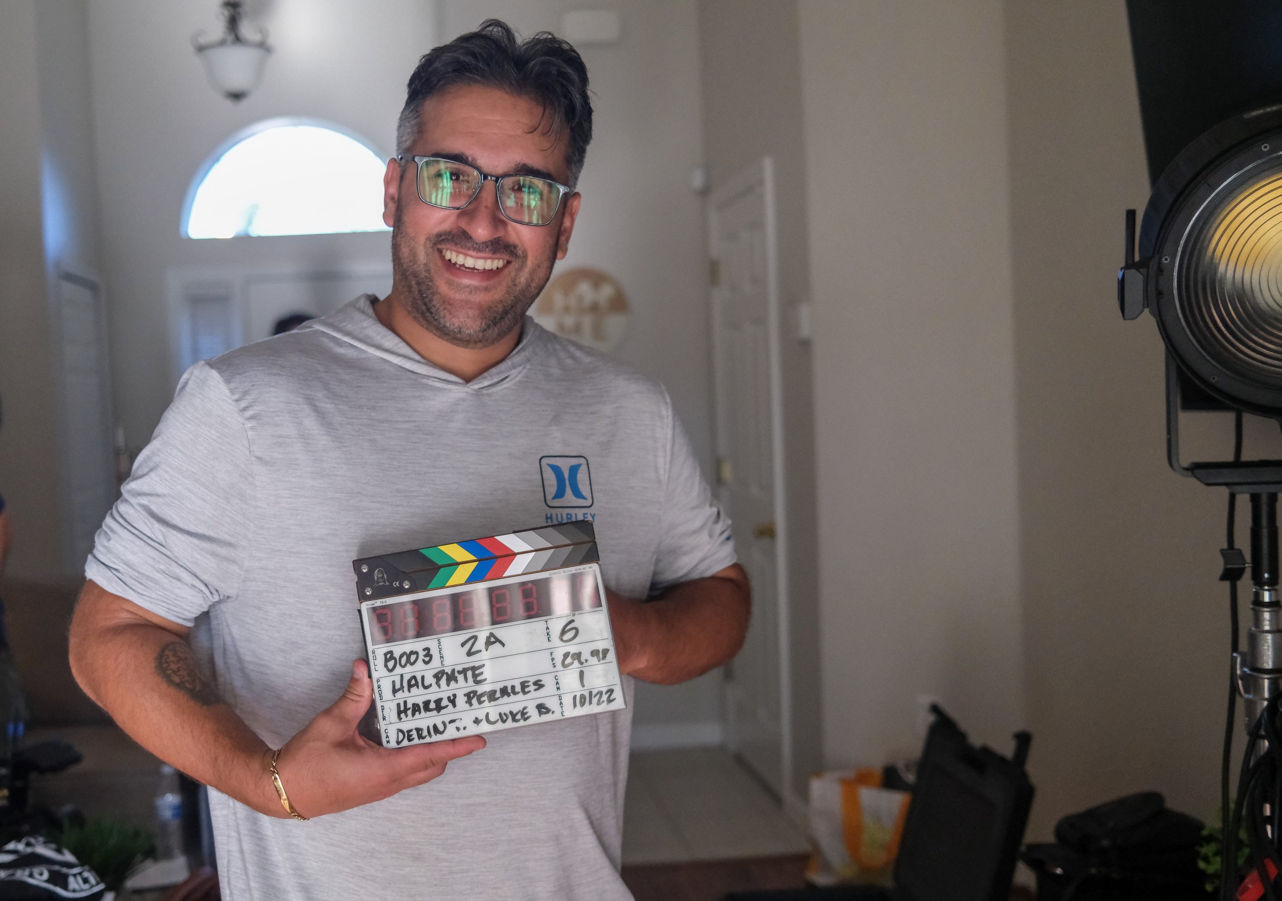 Man with long hair, glasses, and beard smiling, sitting outdoors with film equipment in background.