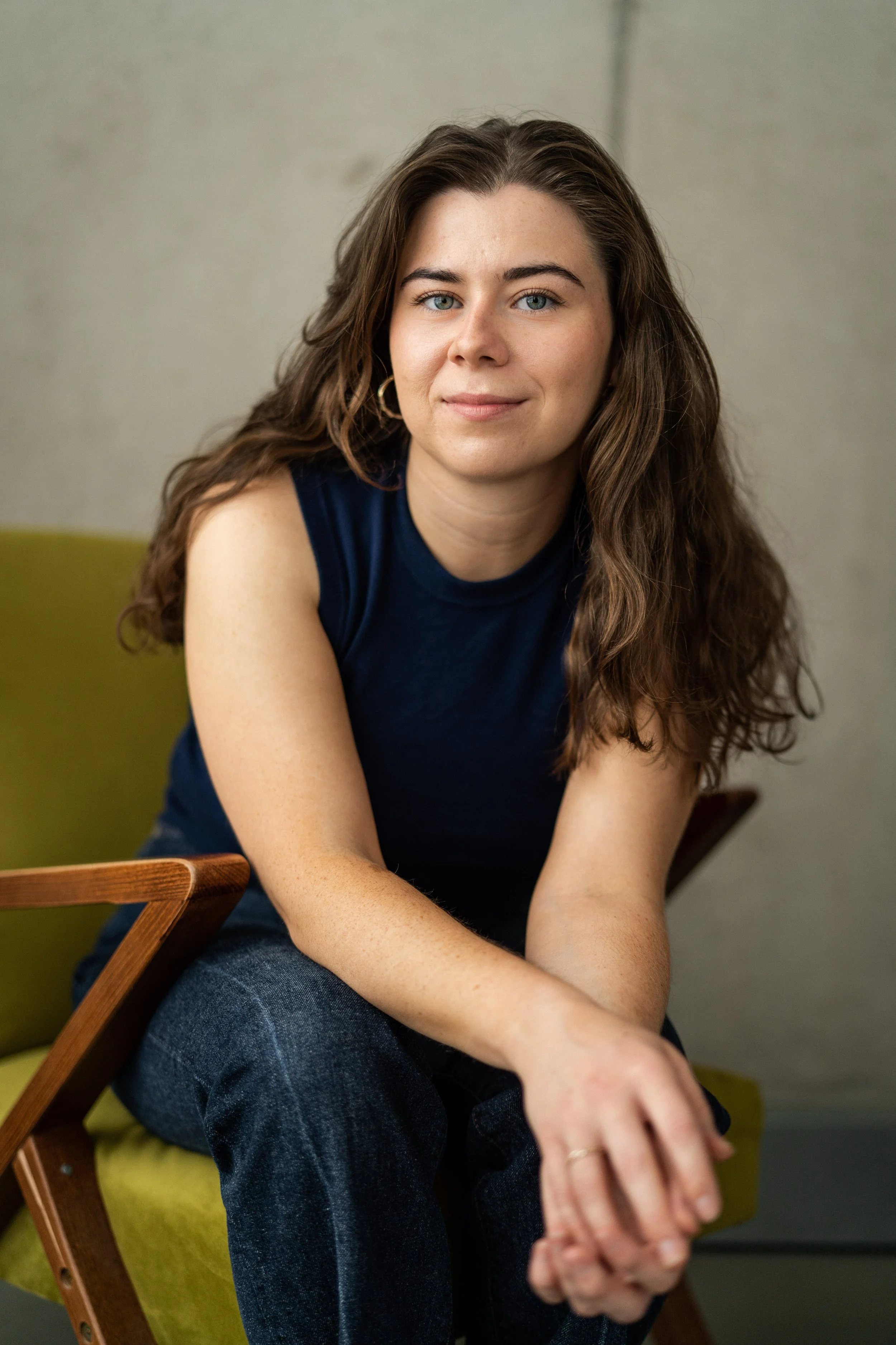 A woman with long, wavy brown hair and blue eyes sitting on a green chair, wearing a sleeveless navy blue top and jeans, looking at the camera and smiling slightly.