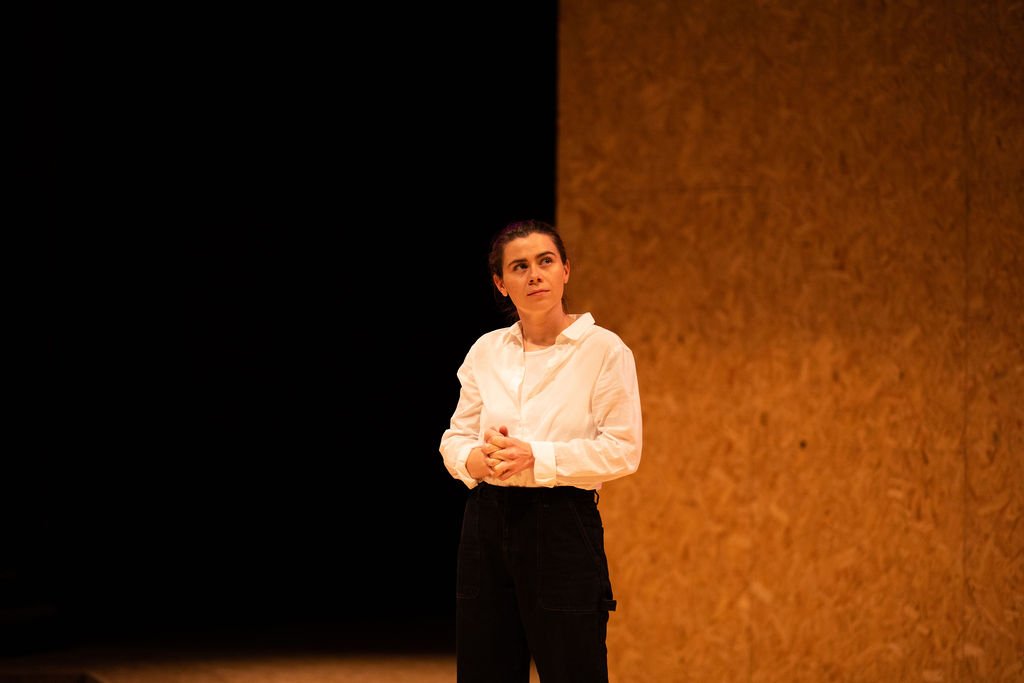 A woman stands on a stage with a wooden background, wearing a white shirt and black pants, looking thoughtful.