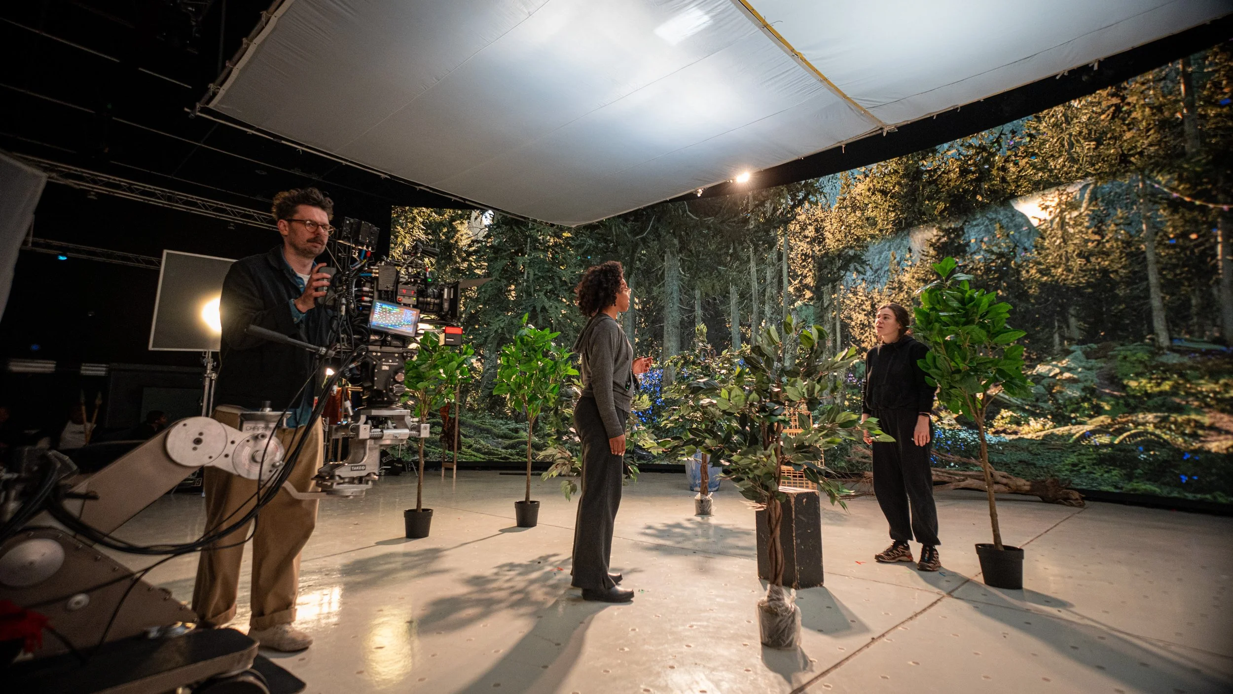 Film crew setting up a scene with two women in a forest background on a green screen, surrounded by potted trees, with a camera operator adjusting a camera and crew members preparing for filming.