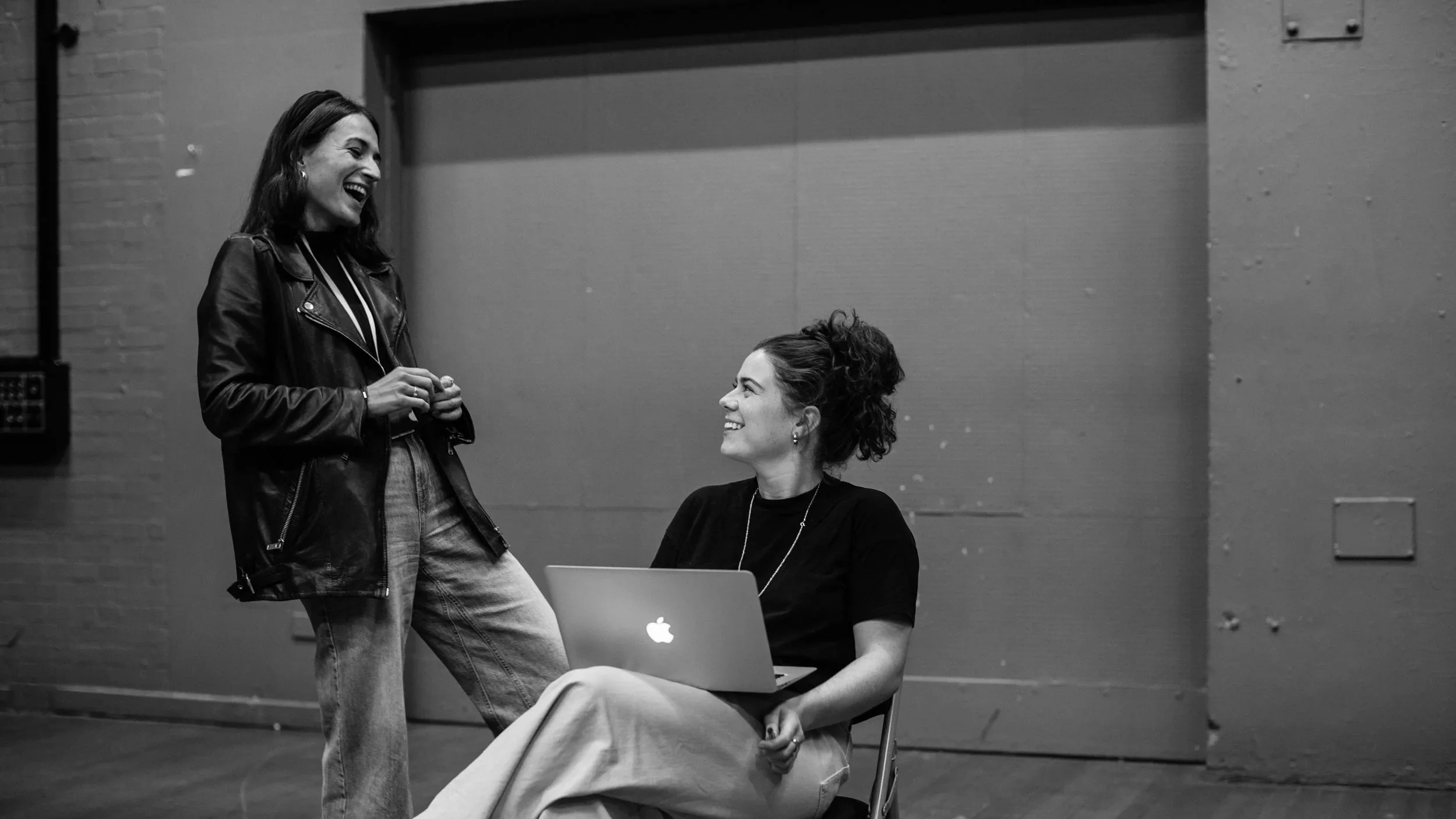 Two women smiling and talking, one sitting with a laptop and the other standing, in an indoor setting.
