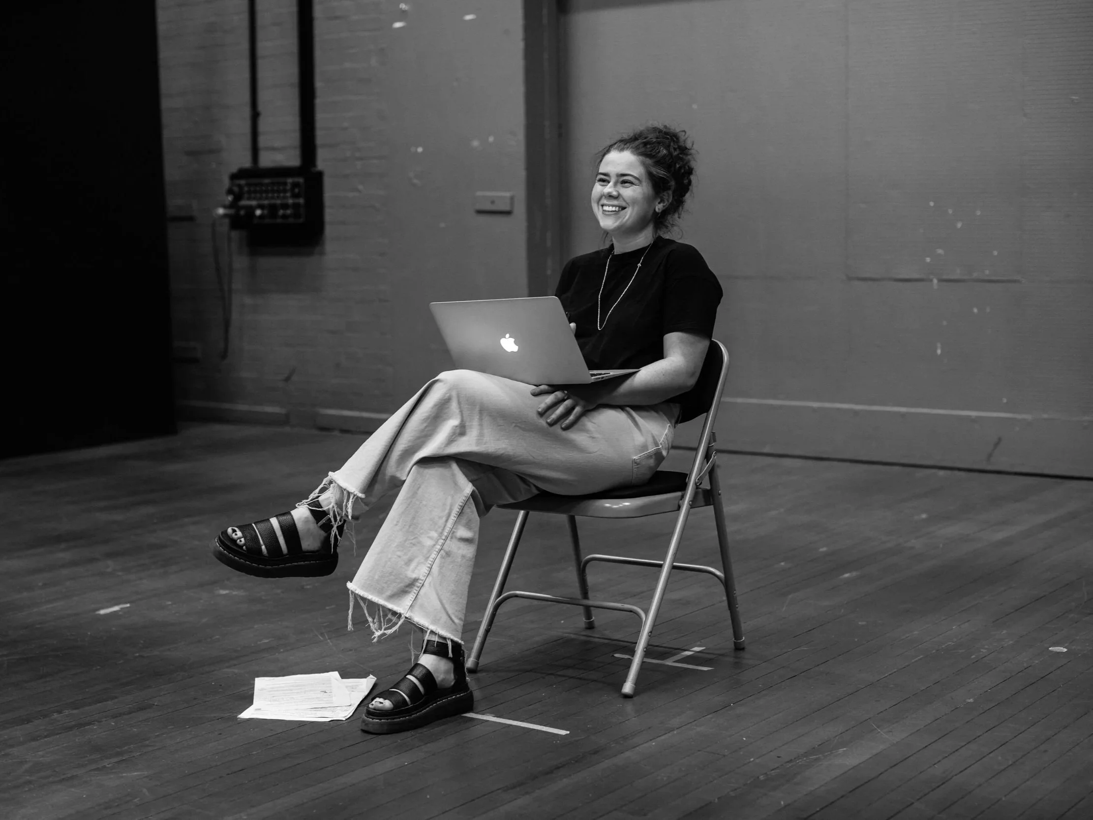 A woman sitting on a chair on a wooden floor, smiling, holding an open MacBook laptop on her lap. She is wearing a black t-shirt, wide-legged ripped jeans, and black sandals with socks. Papers are scattered on the floor beside her. The background features a plain wall and some electrical equipment mounted on it.