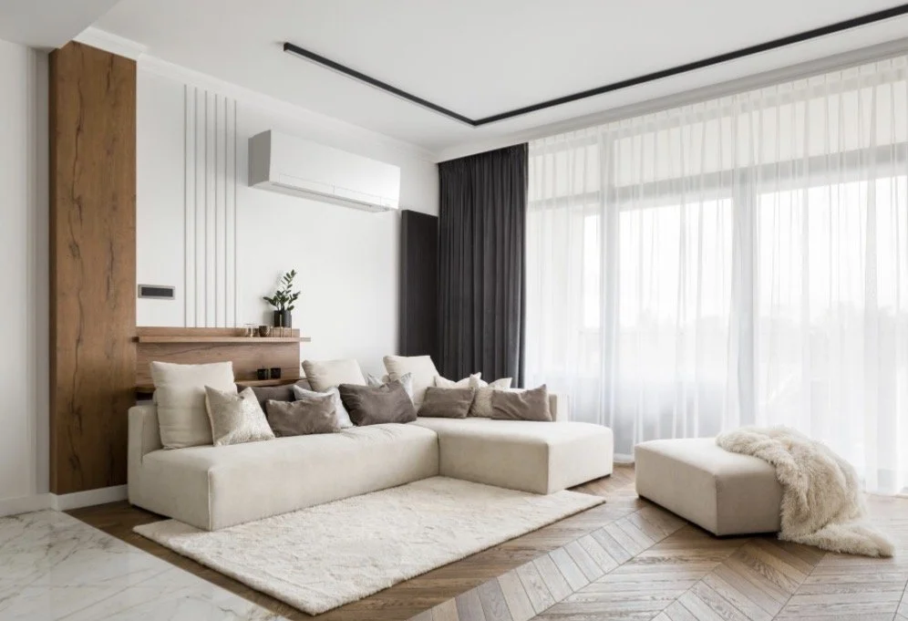 Modern living room with a beige sectional sofa, decorative pillows, a plush rug, sheer curtains, and large windows.