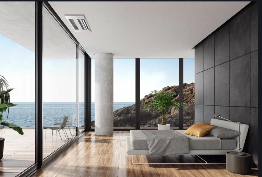 Modern bedroom with large glass windows overlooking the ocean, featuring a white bed with a gray blanket and orange pillow, a potted plant, and a view of a rocky shoreline.