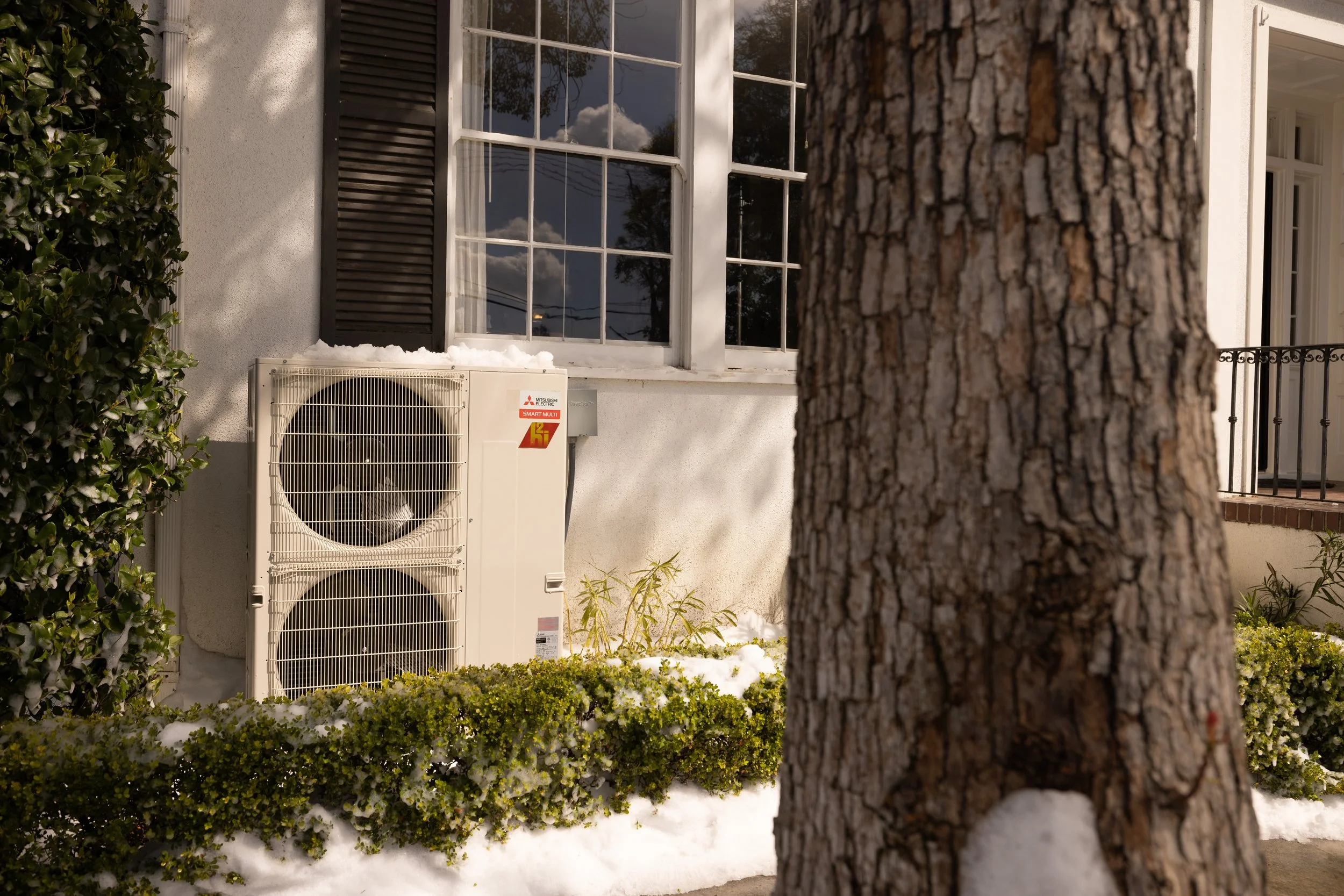 A tree trunk in the foreground, a white air conditioning unit with snow on top, a window with open black shutters, and a porch with a railing in front of a white house with snow on the ground.