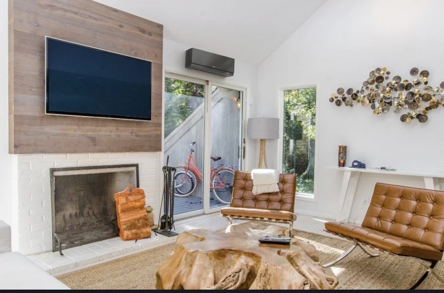 Modern living room with a large wall-mounted TV, a fireplace with a firewood holder, two leather lounge chairs, a floor lamp, and an abstract metal wall art piece. Large glass sliding doors lead outside, revealing bicycles on a patio.