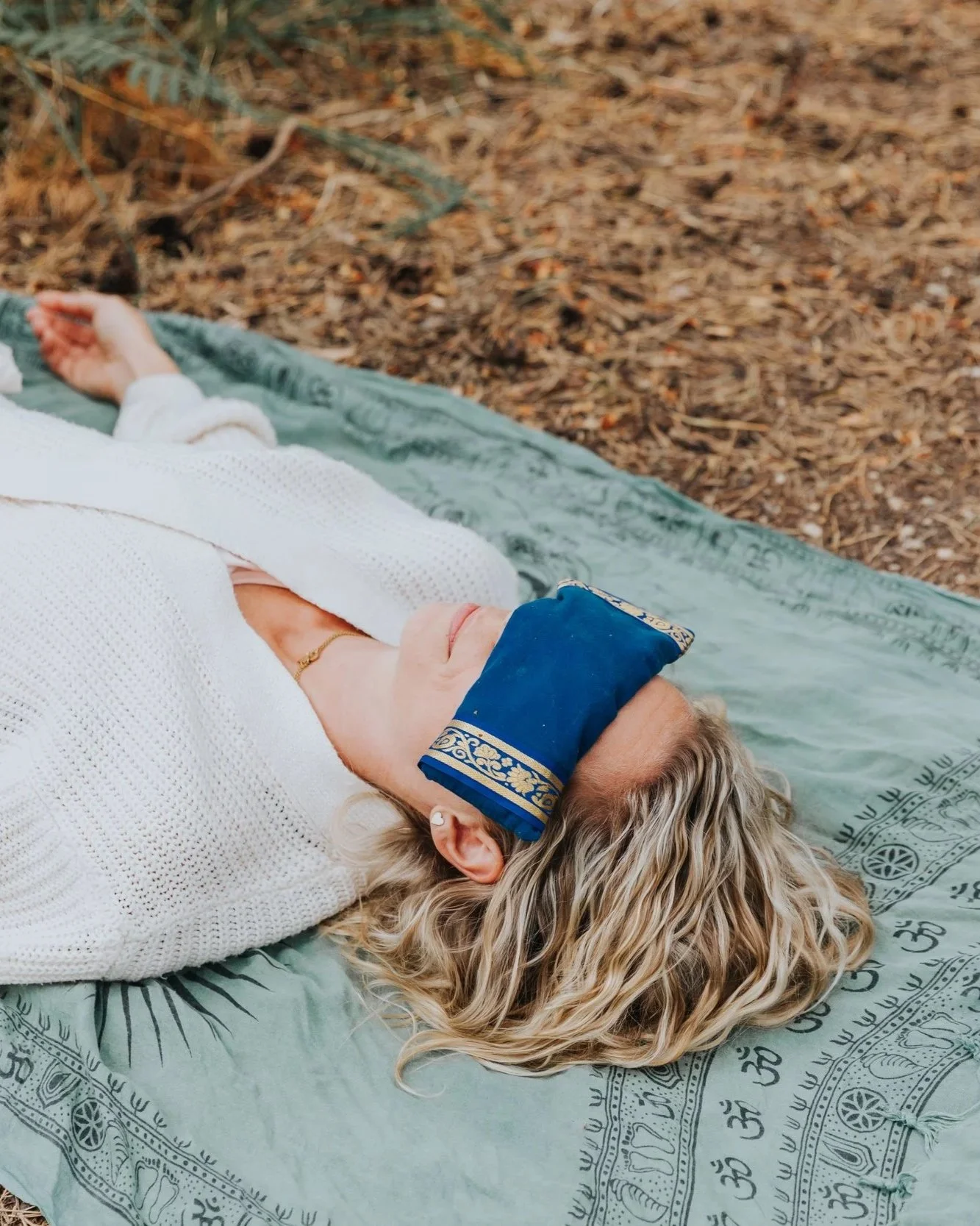 A woman with blonde hair relaxing outdoors on a green blanket with spiritual symbols, wearing a blue prayer shawl over her eyes and a white sweater, surrounded by natural forest setting.