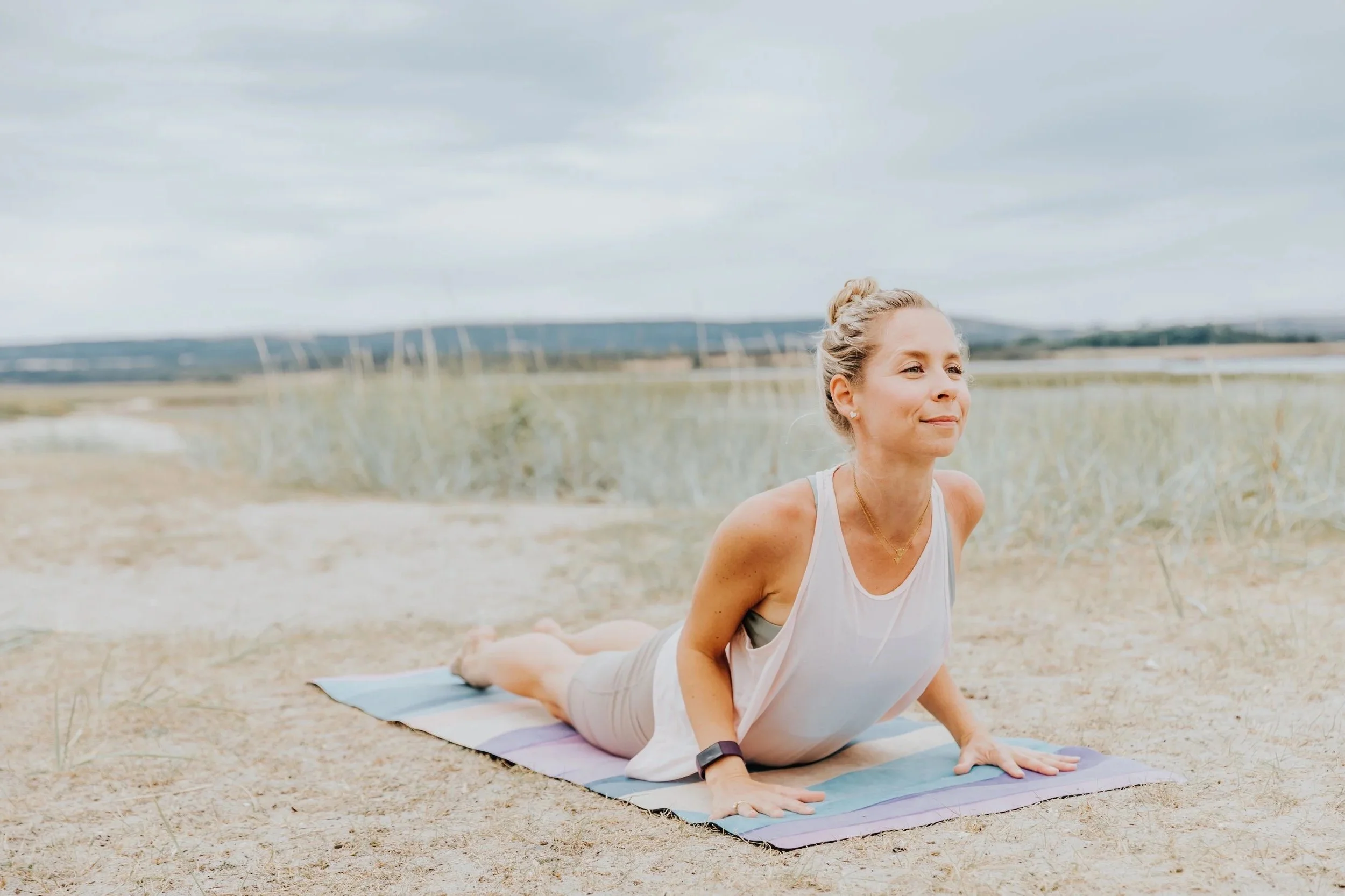 A woman practicing yoga outdoors on a beach, doing a cobra pose on a yoga mat with a scenic landscape of water and hills in the background.