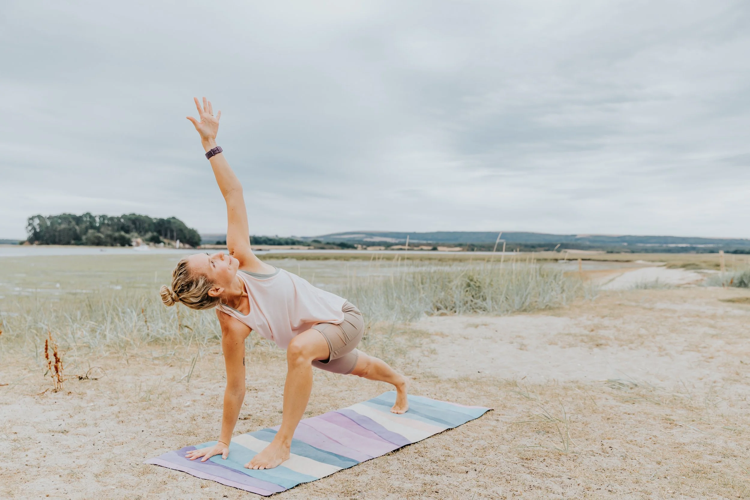 A woman practicing yoga outdoors on a striped mat near a sandy beach with grassy dunes, exercising a side plank pose with one arm extended upward on a cloudy day.