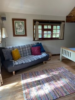 A cozy room with a gray plaid couch accented by yellow and purple pillows, a colorful patterned rug, a window with curtains, a framed artwork on the wall, and a white crib in the corner.