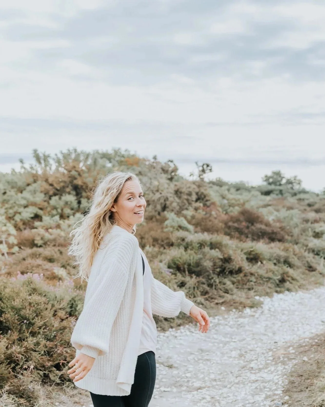 A woman with long blonde hair walks along a rocky path through a desert-like landscape with bushes and shrubs, under a cloudy sky.