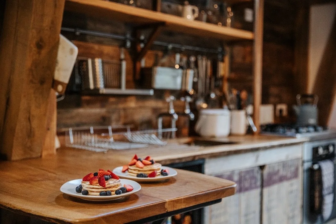 Two plates of pancakes topped with strawberries and blueberries on a wooden countertop in a rustic kitchen.