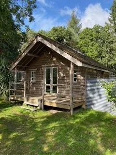A small wooden cabin with a front porch, set in a lush green area with trees and a partly cloudy sky.