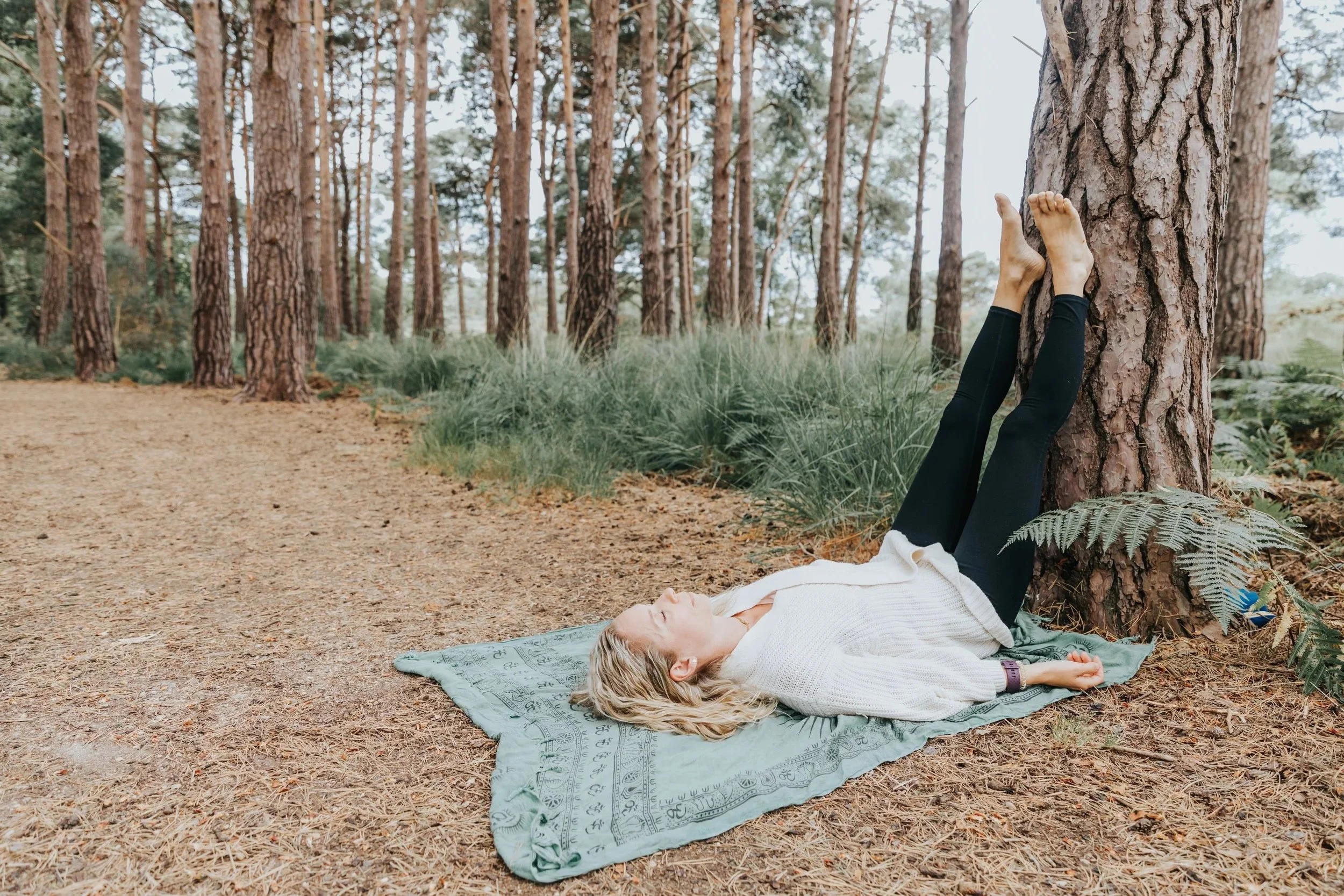 A woman practicing yoga or stretching in a forest, lying on a blanket with her legs against a tree trunk.