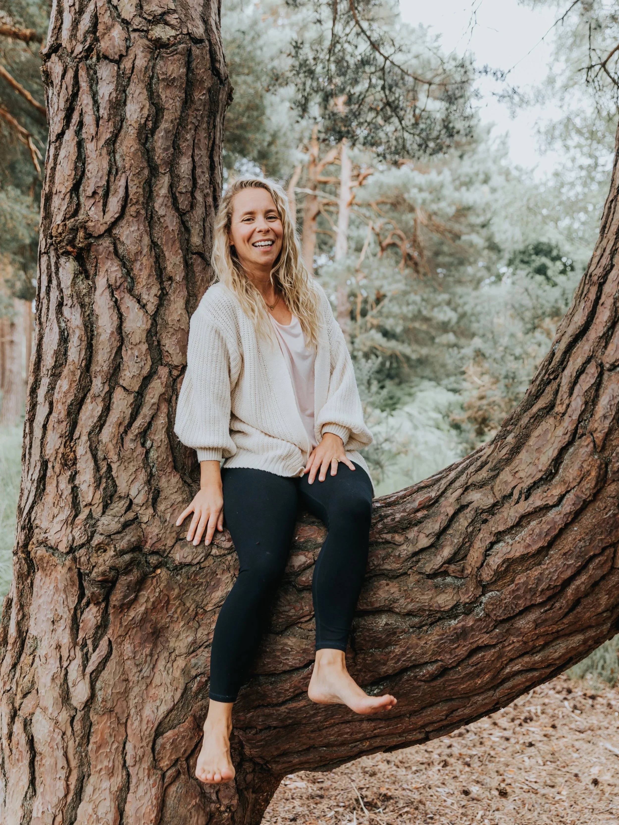 A woman with blonde, curly hair smiling while sitting on a large tree branch in a forest, wearing a cream-colored sweater and black leggings.
