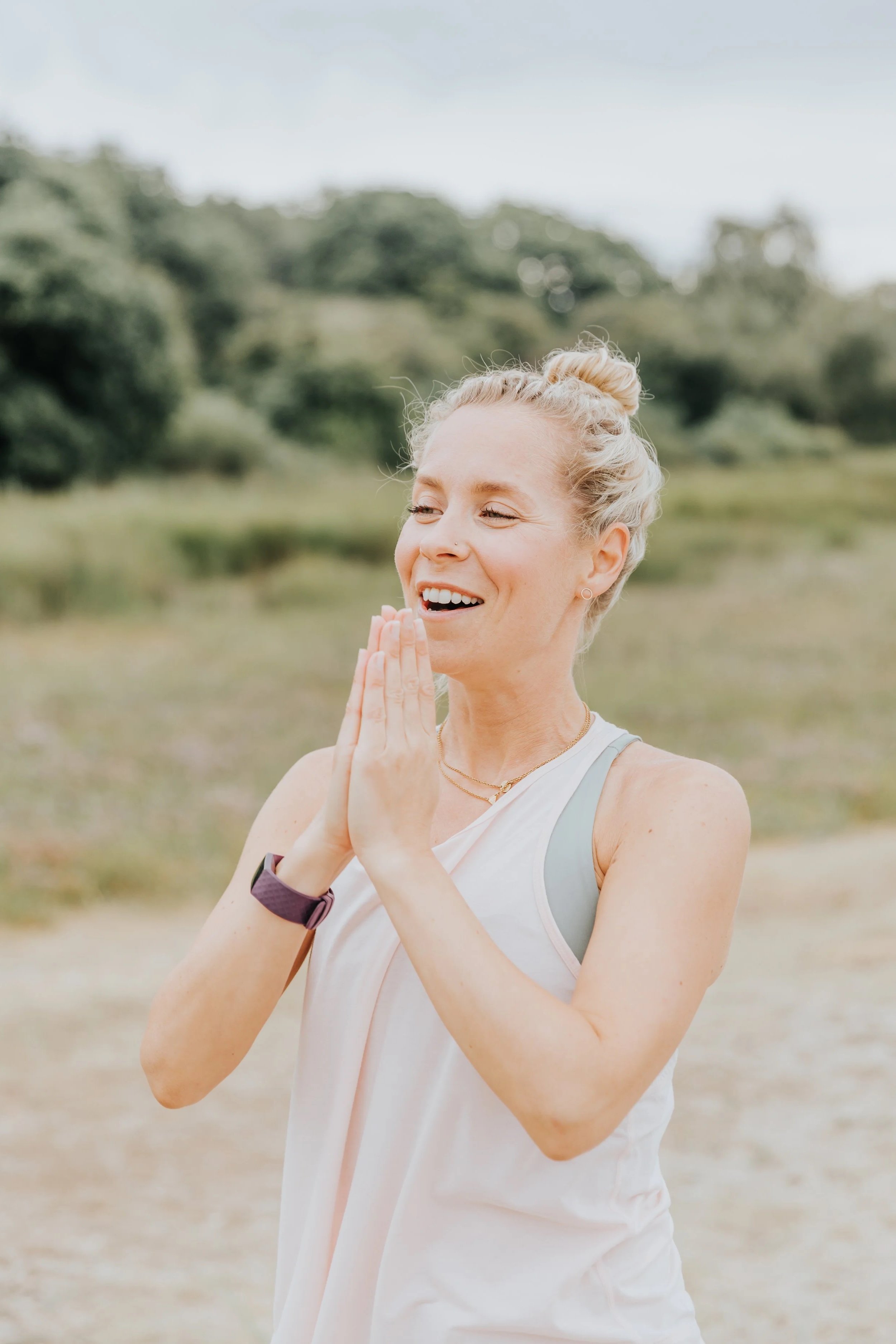 A woman with blonde hair in a bun, wearing a white tank top, smiling with her eyes closed, and hands pressed together in front of her chest, standing outdoors near water and trees.