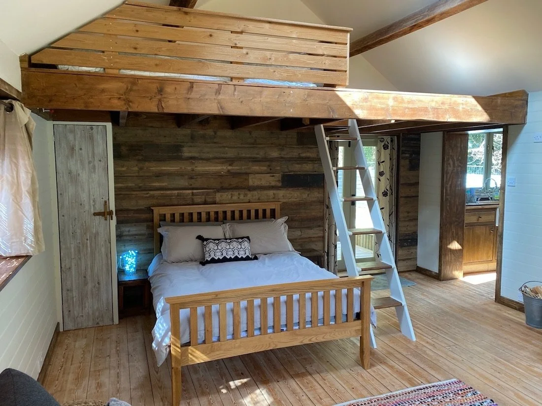 A cozy bedroom with a wooden bed, wooden wall, and a loft area accessible by a ladder. The room has wooden floors, a window with curtains, and is adjacent to a small kitchen area.