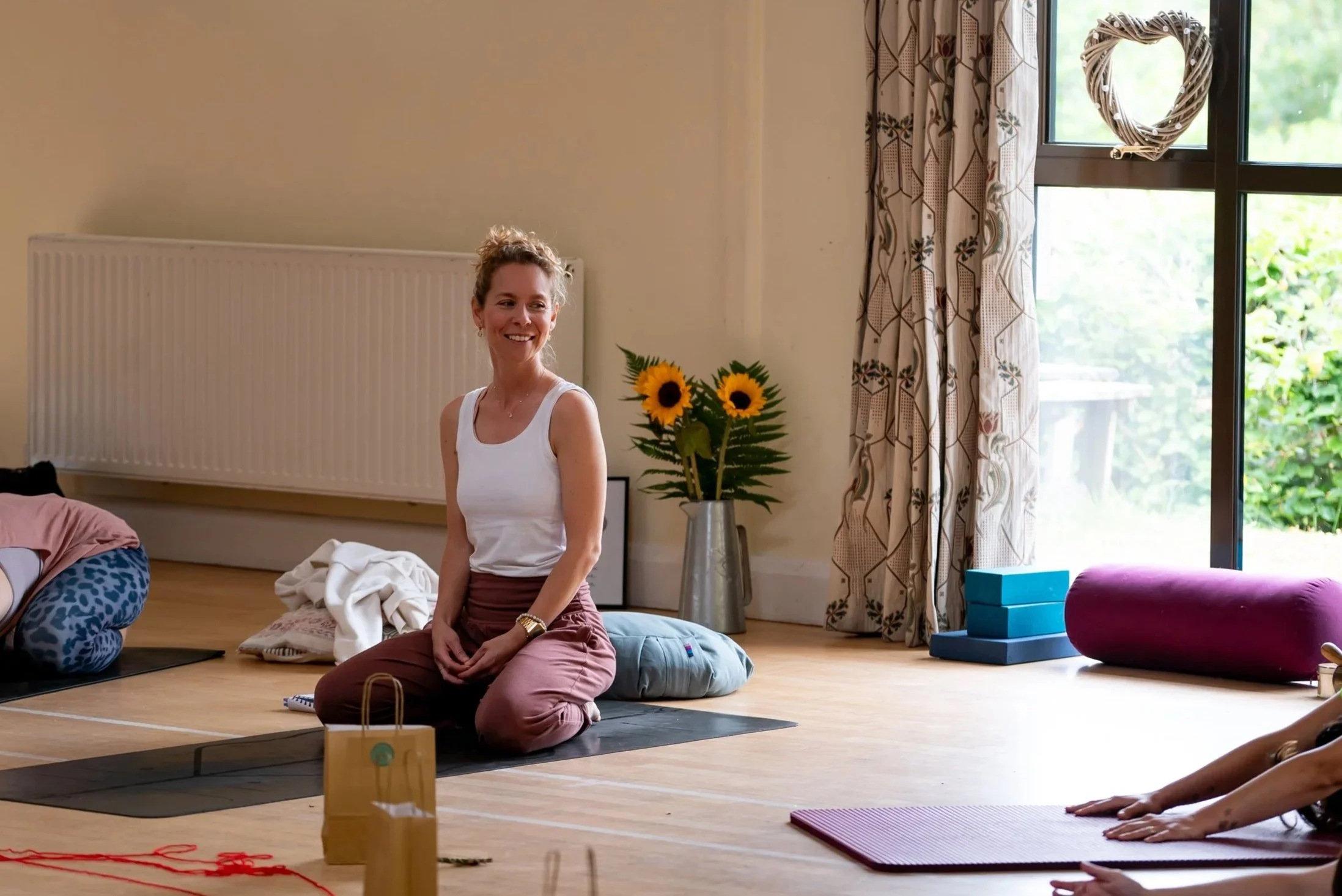 A woman smiling while kneeling on a yoga mat during a yoga class in a bright, cheerful room with large windows, floral curtains, and a vase of sunflowers.