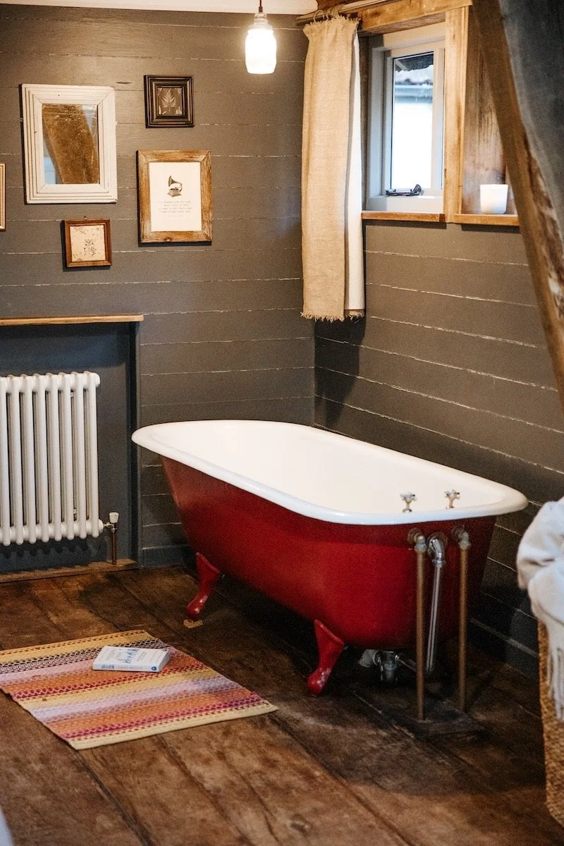 A vintage-style bathroom corner with a red clawfoot bathtub, wooden wall panels, a small window with beige curtains, a white radiator, a hanging light fixture, a multi-colored striped rug, and framed pictures on the wall.