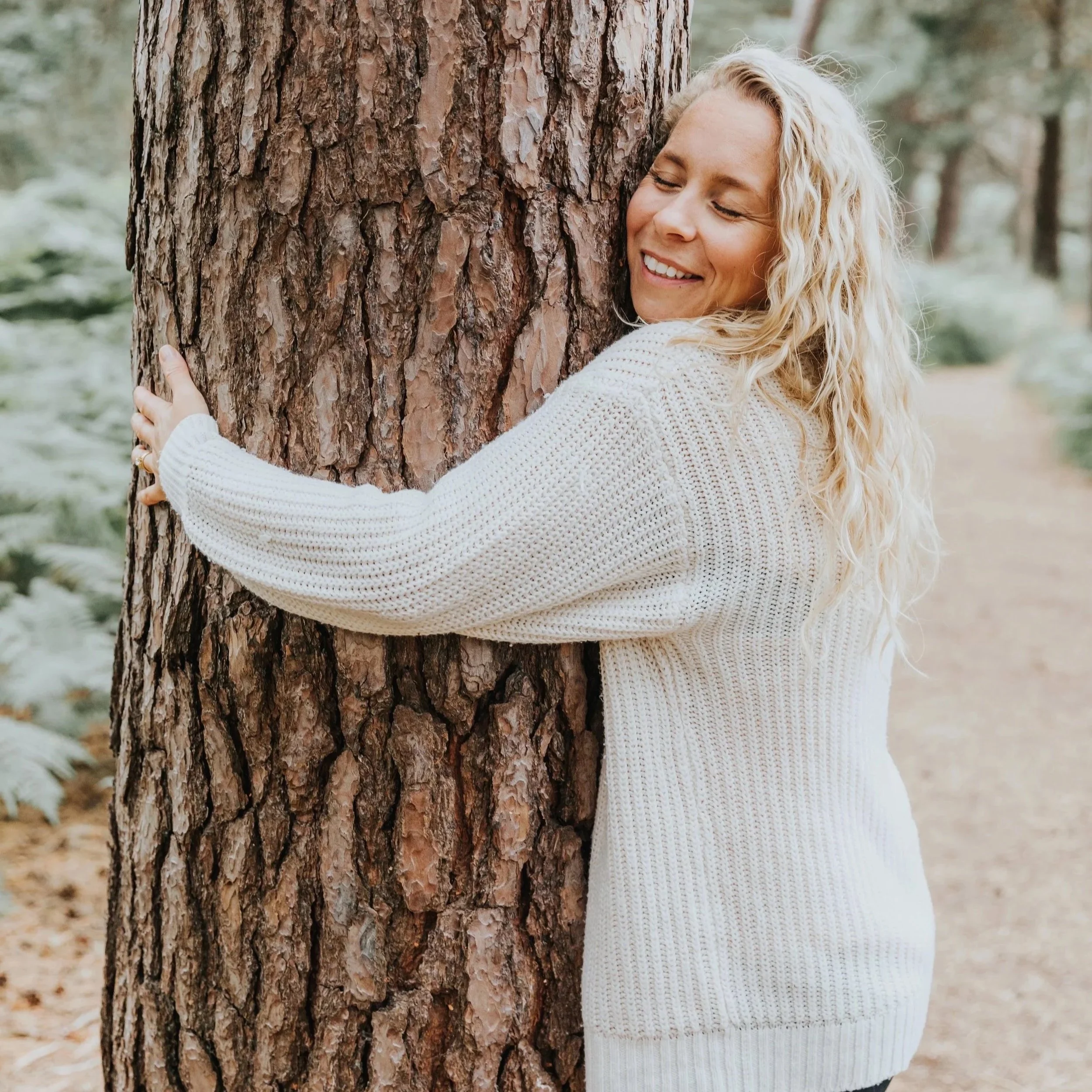 A woman with curly blonde hair hugging a large tree trunk in a forest, smiling with eyes closed.