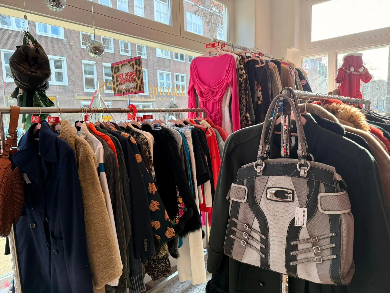 Inside a vintage clothing store with racks of colorful jackets, shirts, and dresses, and a window view of a city street outside. Secondhand vintage tweedehands kleding kinderen volwassenen adults kids