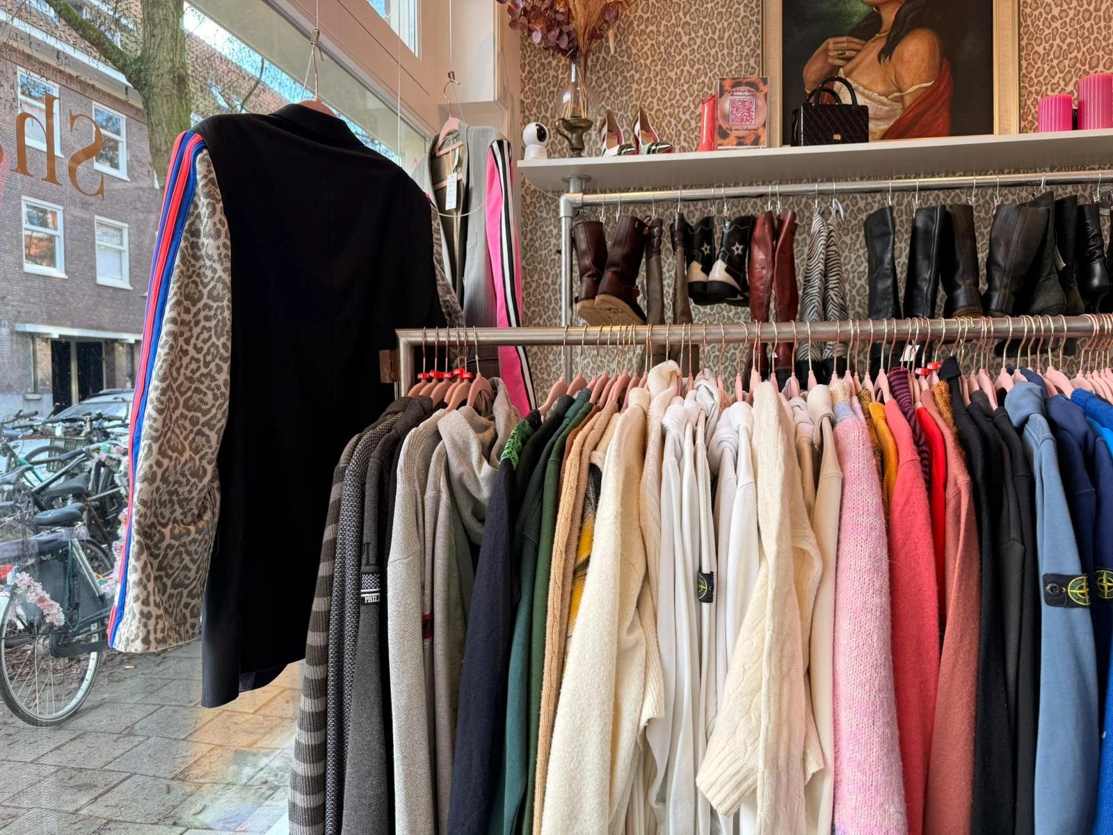 Clothing store window display with jackets, hoodies, and boots on hangers and shelves, looking out onto a city street with bicycles outside.