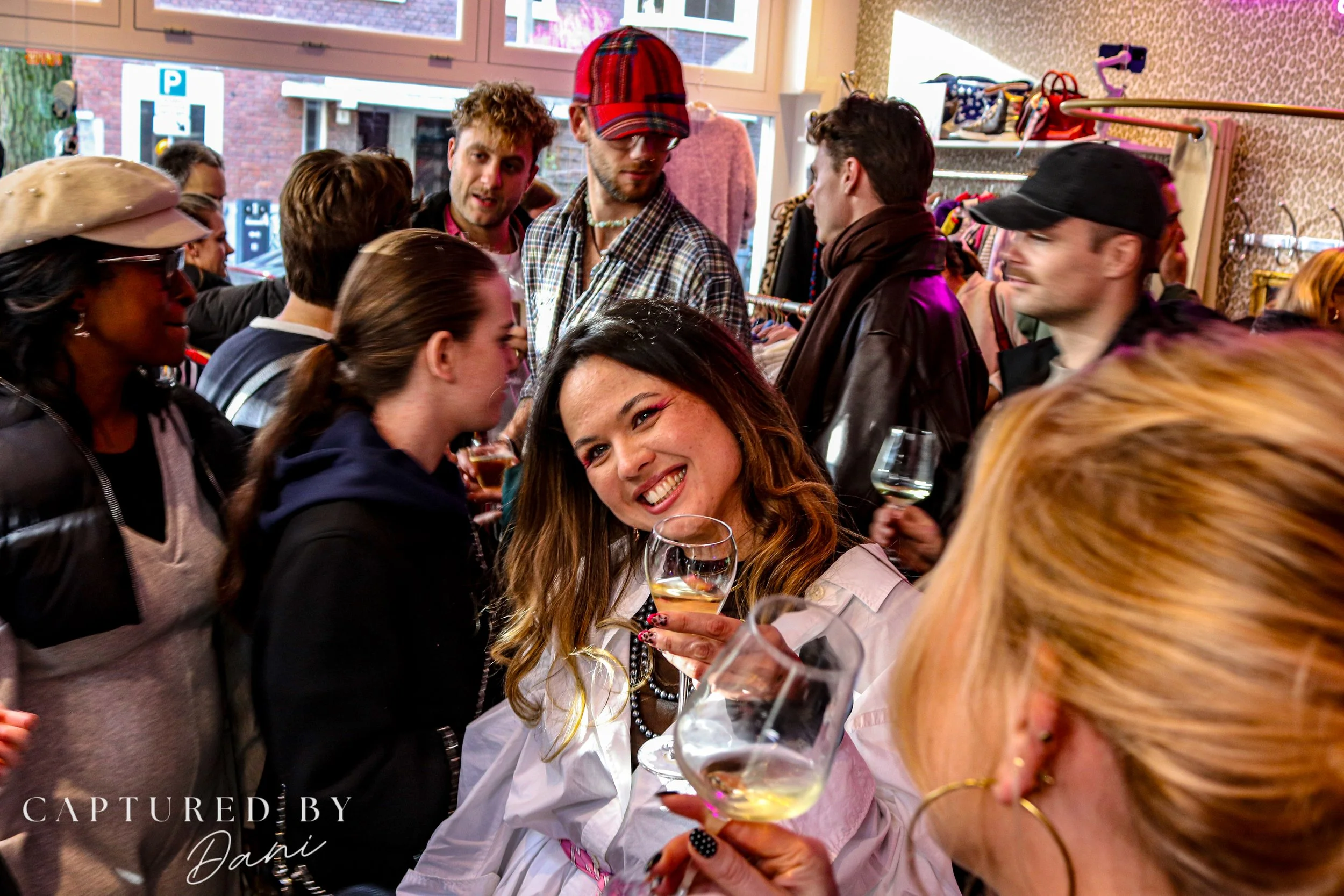 A woman with long wavy hair smiling and holding a glass of white wine at a lively social gathering or party with other people, some holding glasses of wine and engaging in conversation inside a well-lit room.