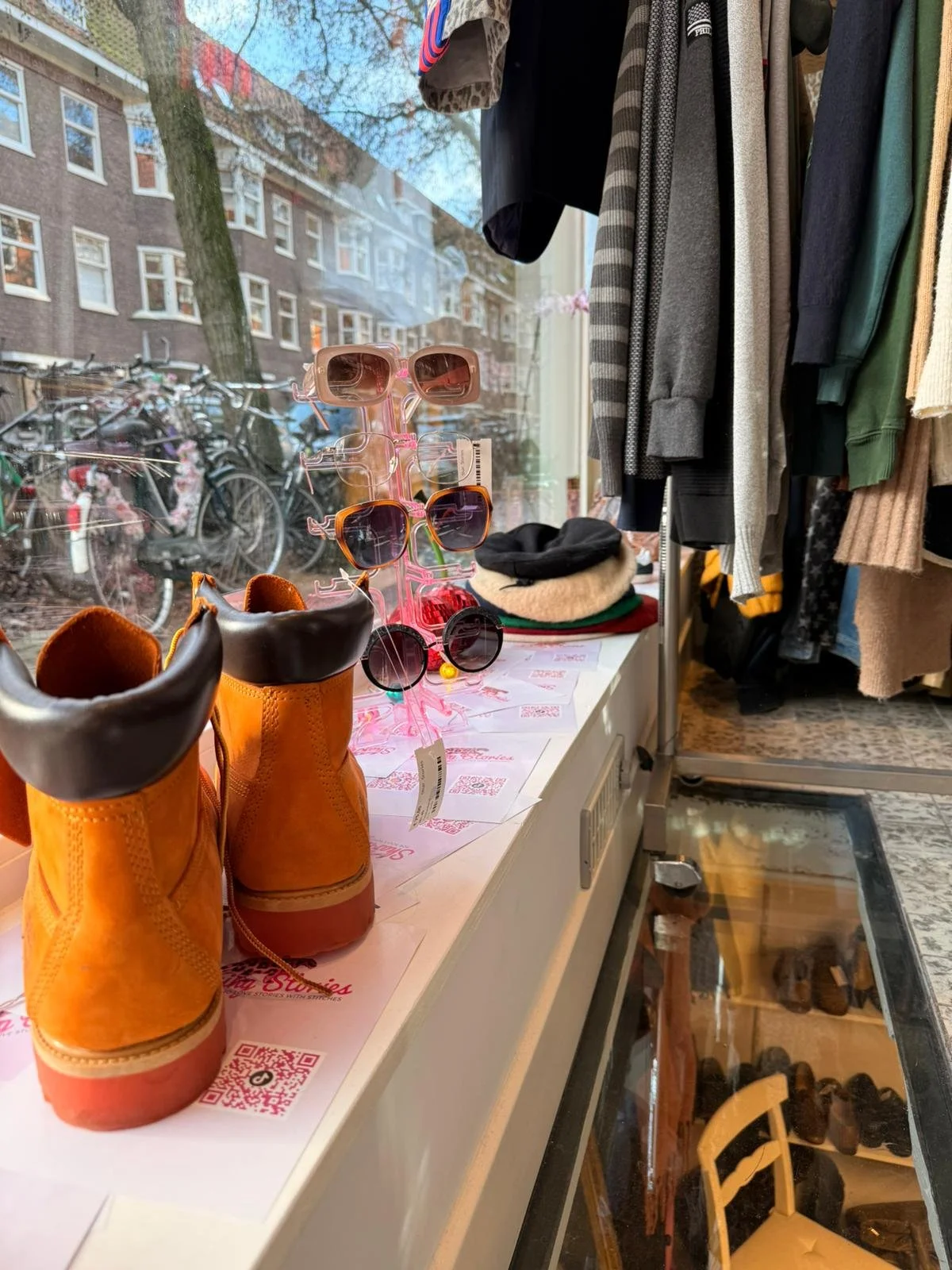 A display of sunglasses on a pink stand, with Timerland boots in front, inside a thrift shop. Kleding hangt hier in de ramen, kleurrijk en met prints. Tweedehands secondhand