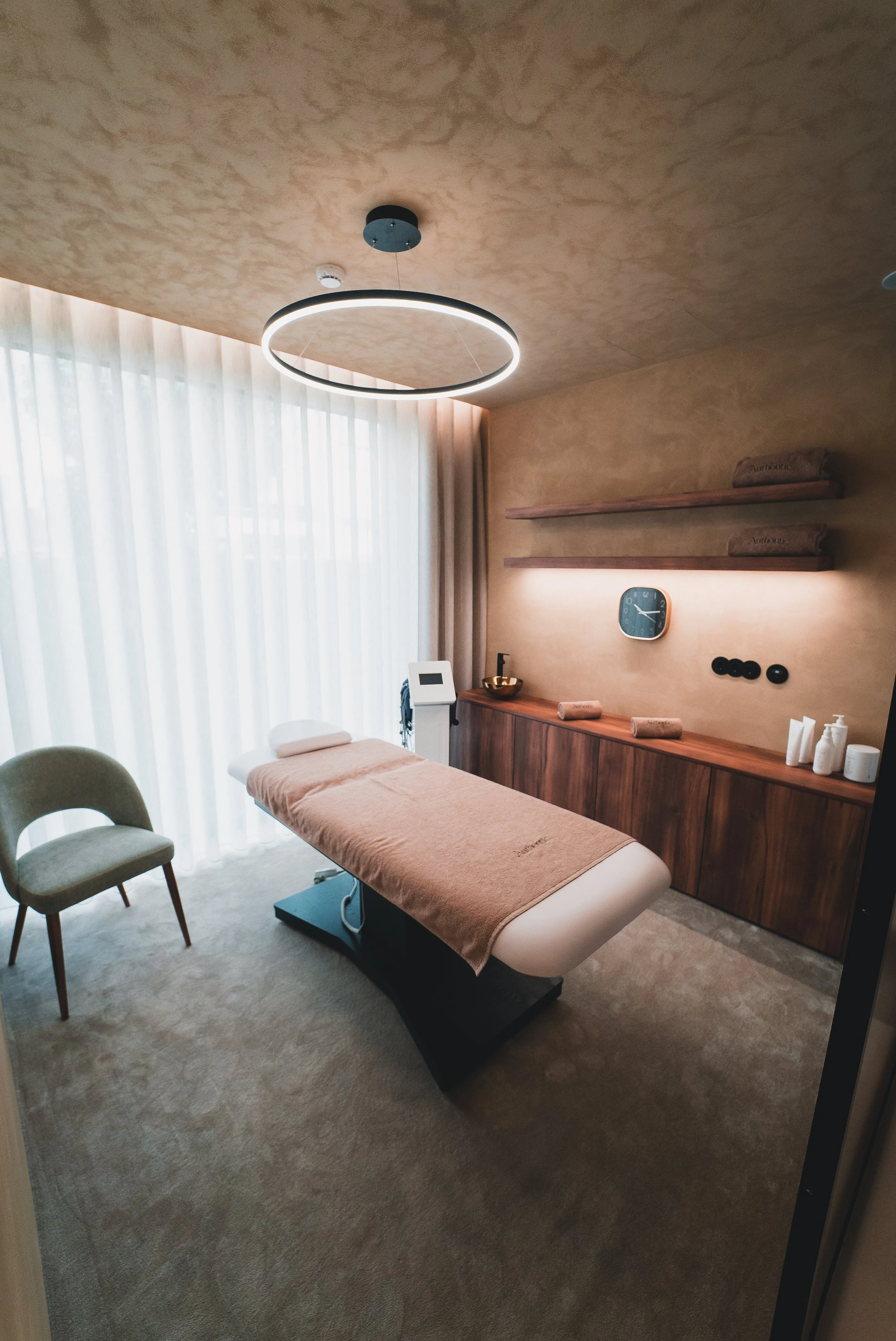 Sala de terapia com uma mesa de massagem, cadeira, cortinas de tecido e iluminação moderna no teto.