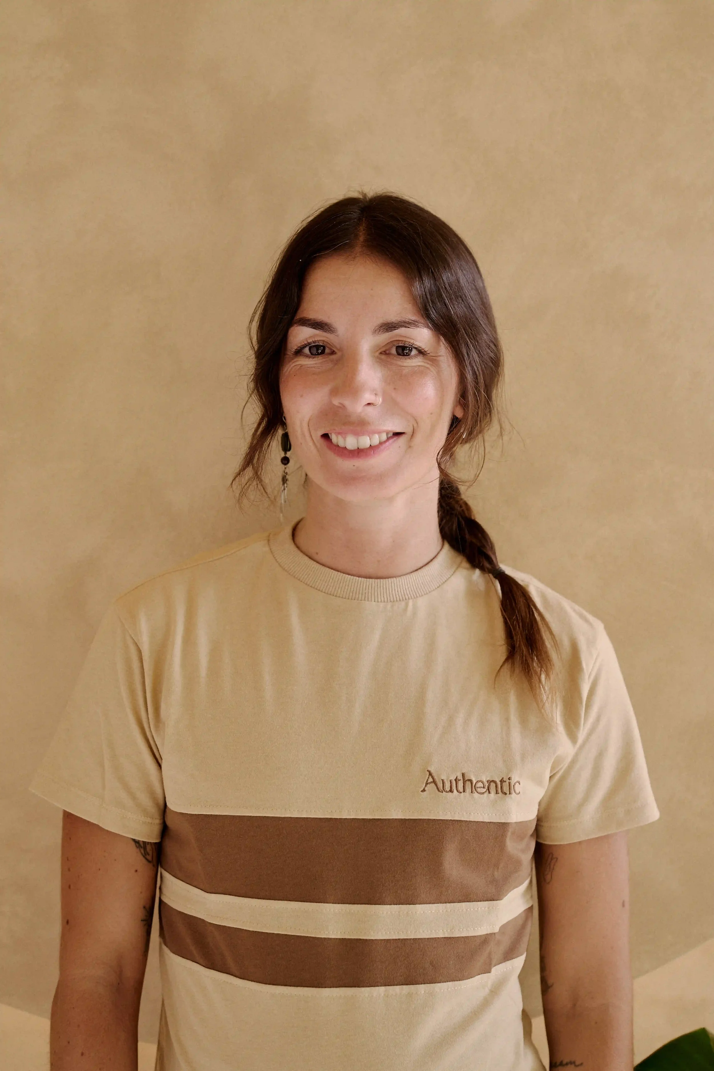 Mulher Professora de Barre e Yoga e Pilates do Authentic Premium Studio, usando camiseta bege com faixas marrons e creme, de brincos pequenos, posando contra parede bege.