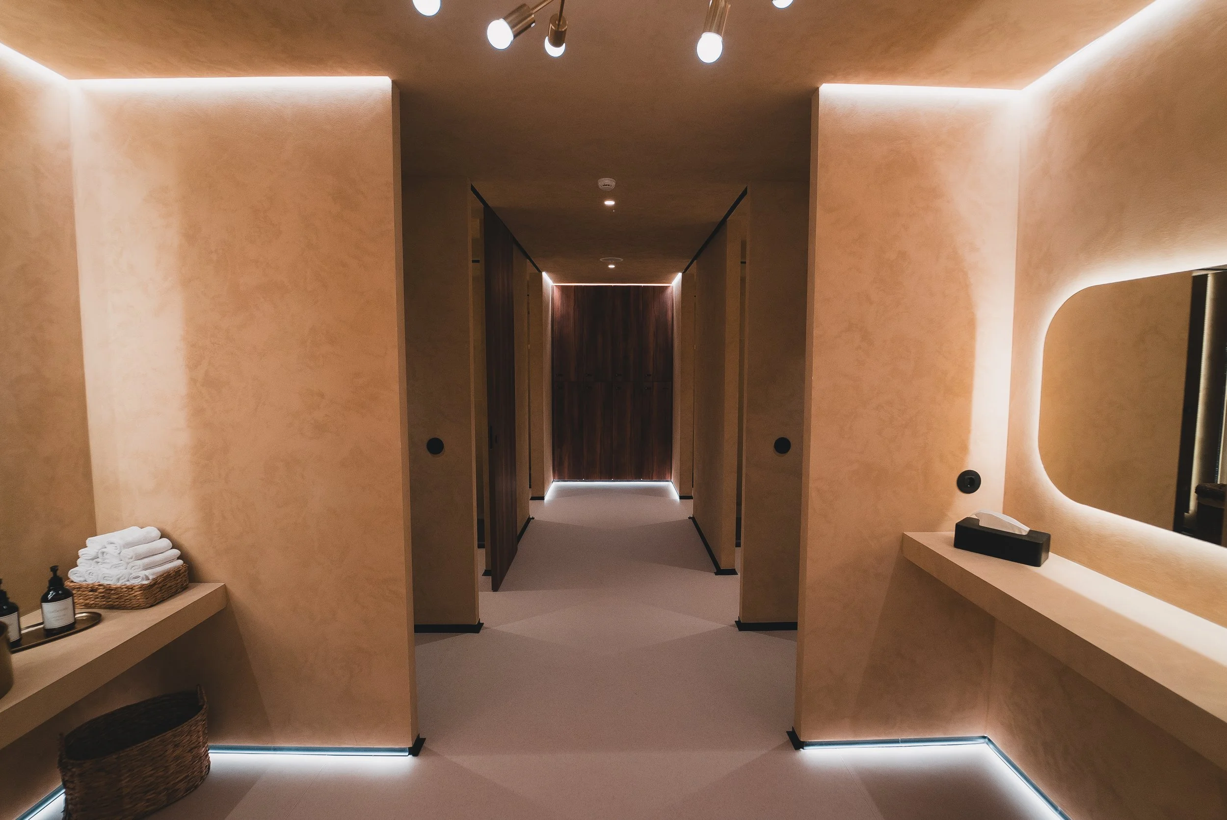 Corredor com paredes de tom bege, iluminação indireta no teto e no chão, portas de madeira ao fundo, espelho com moldura arredondada na parede à direita, mesa com caixa de lenços e bandeja com produtos no lado direito, e cesto de roupa na prateleira 