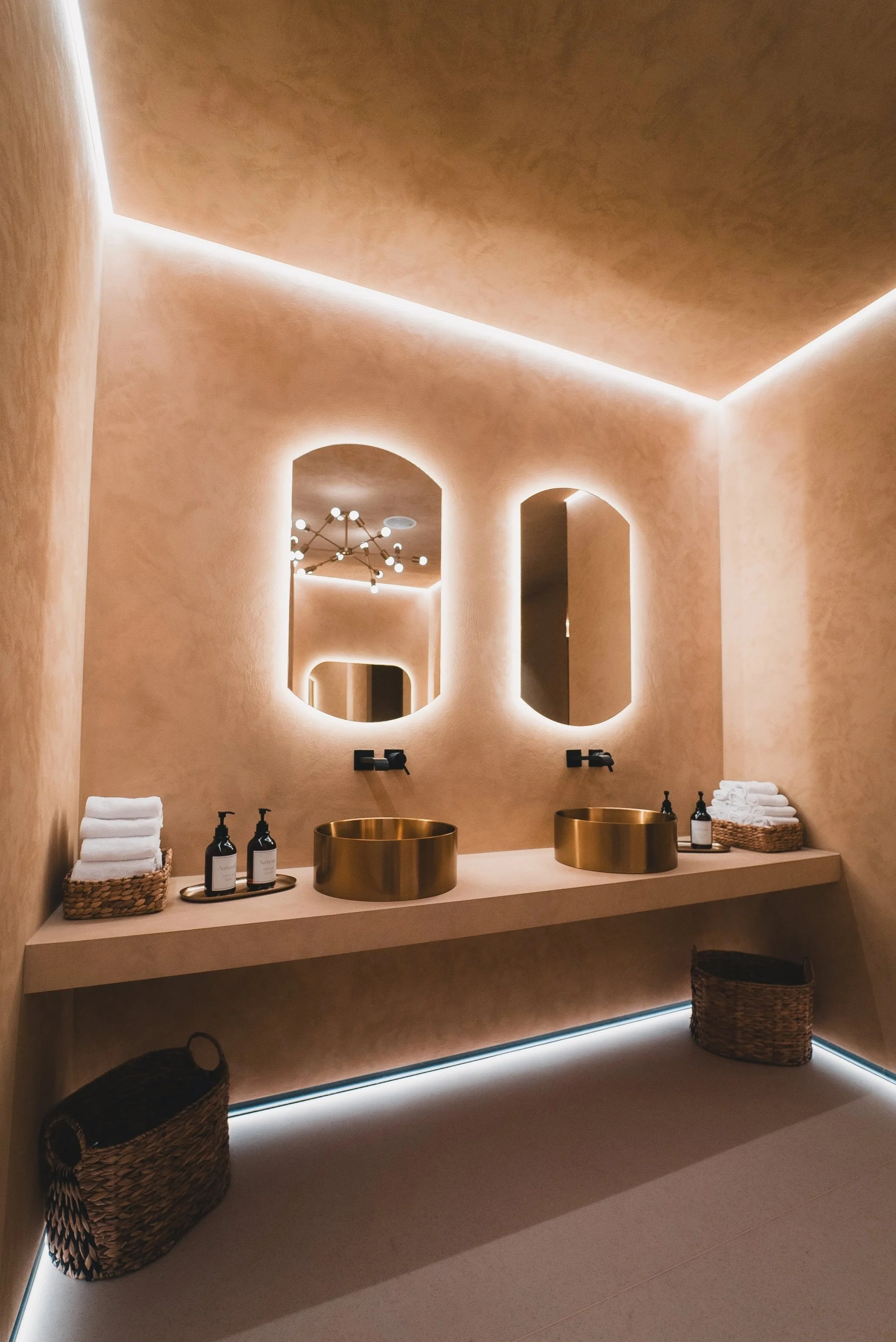 Banheiro com paredes em tom bege, duas torneiras pretas, duas pias douradas em formato de tigela, espelhos retangulares com iluminação ao redor, cestas de vime com toalhas brancas, iluminação de LED na parte inferior do balcão e no teto, e um lustre 