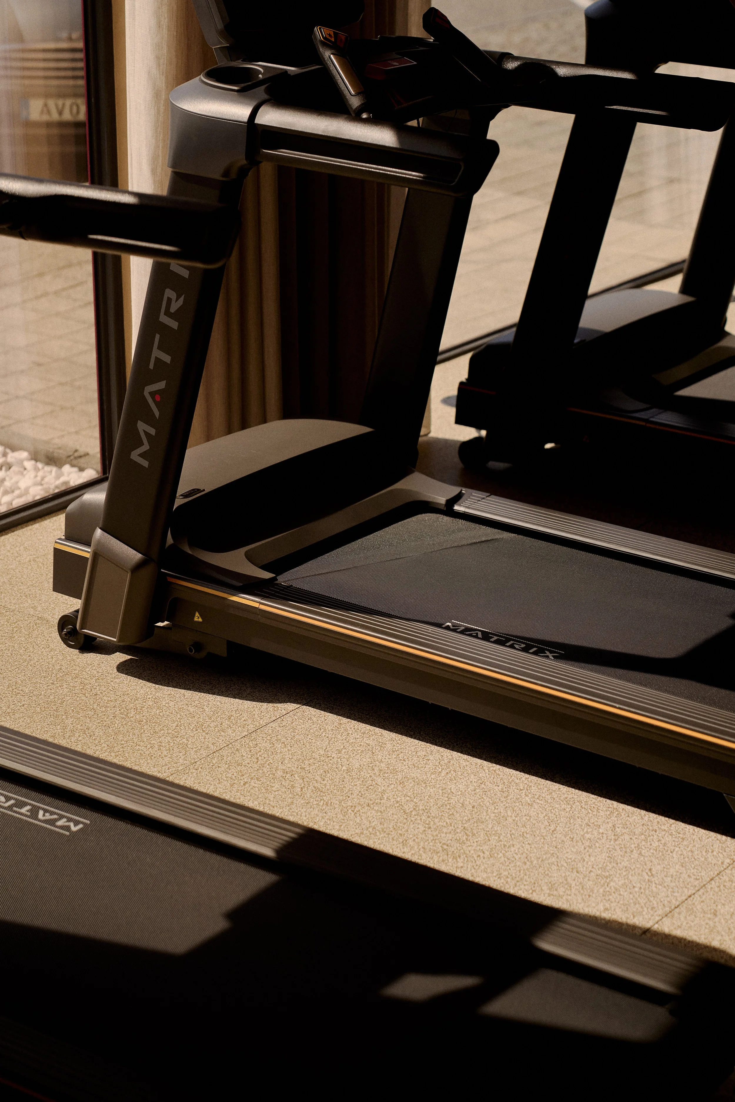 Este é um esteira de corrida preta com detalhes de cor dourada, localizada perto de uma janela com luz natural entrando.