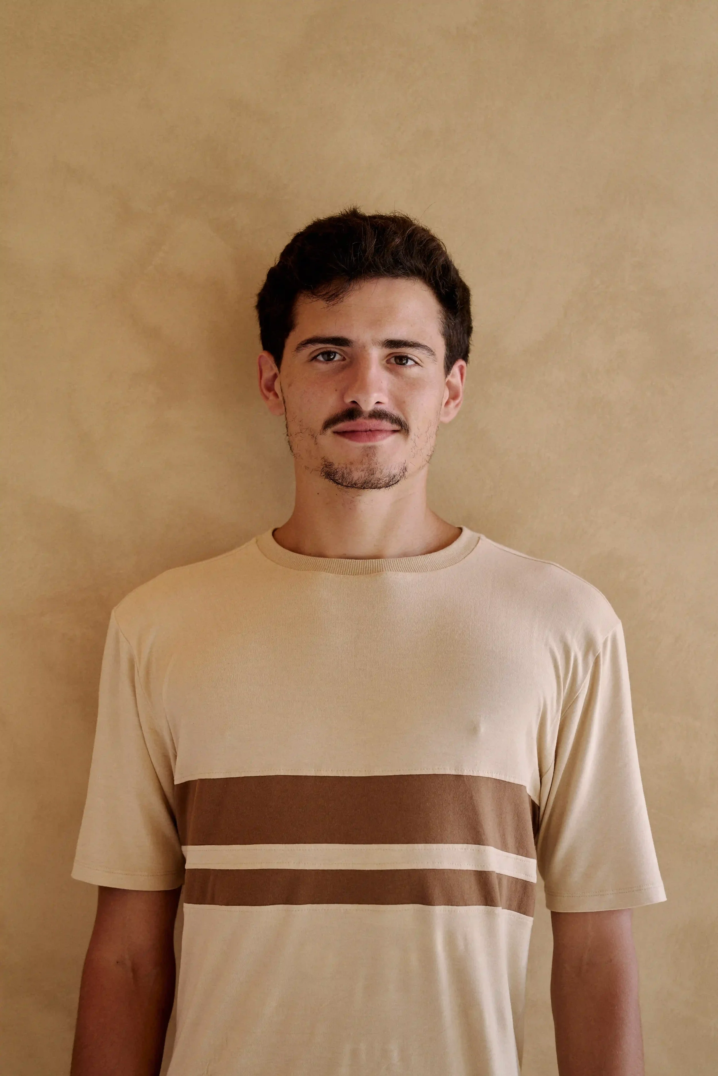 Homem Personal Trainer do Authentic Premium Studio, vestindo camiseta bege com faixas marrons, olhando para a câmera, parado contra uma parede bege.