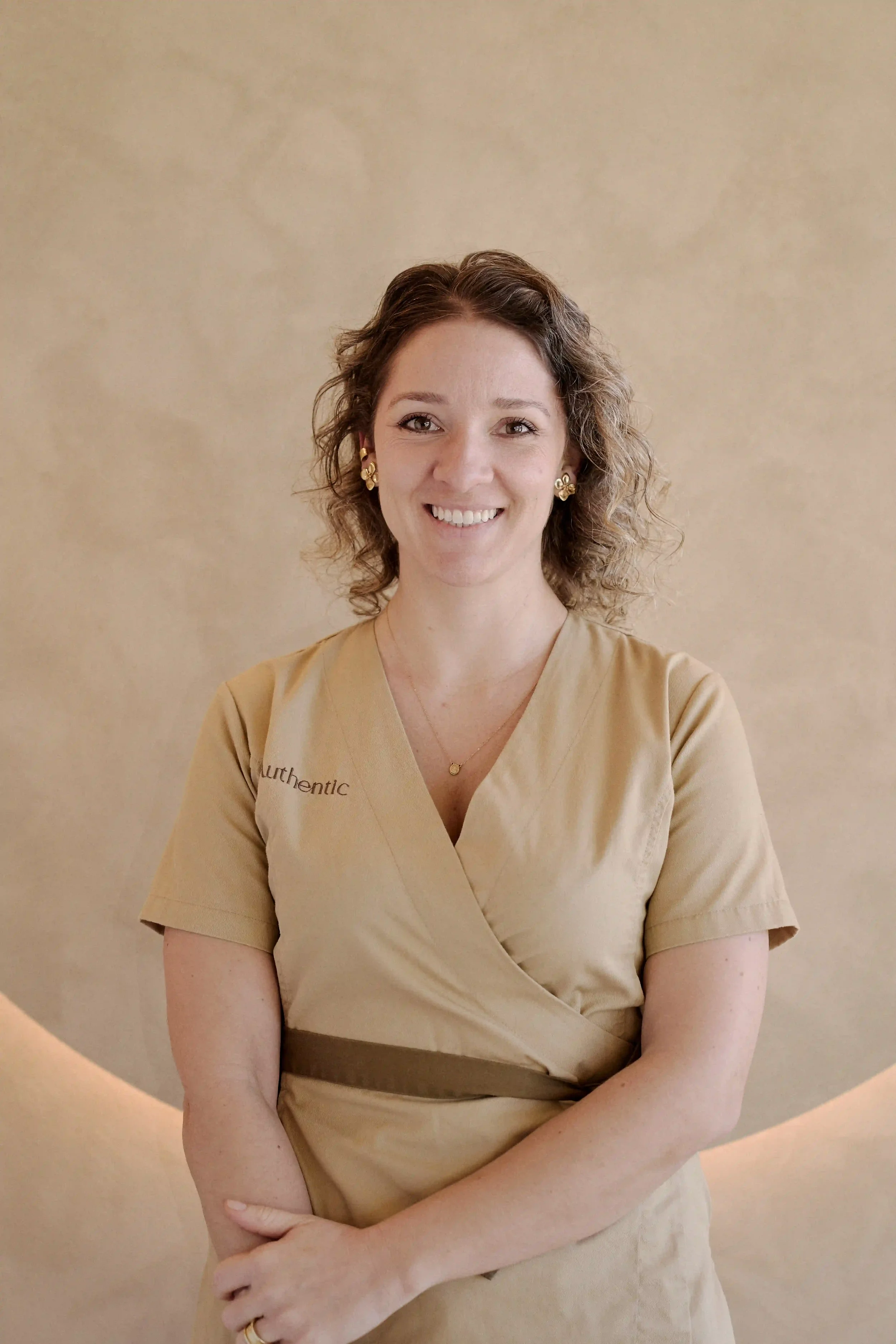 Mulher a sorrir com casaco beje com a palavra "Authentic" escrito, parada em frente a parede bege