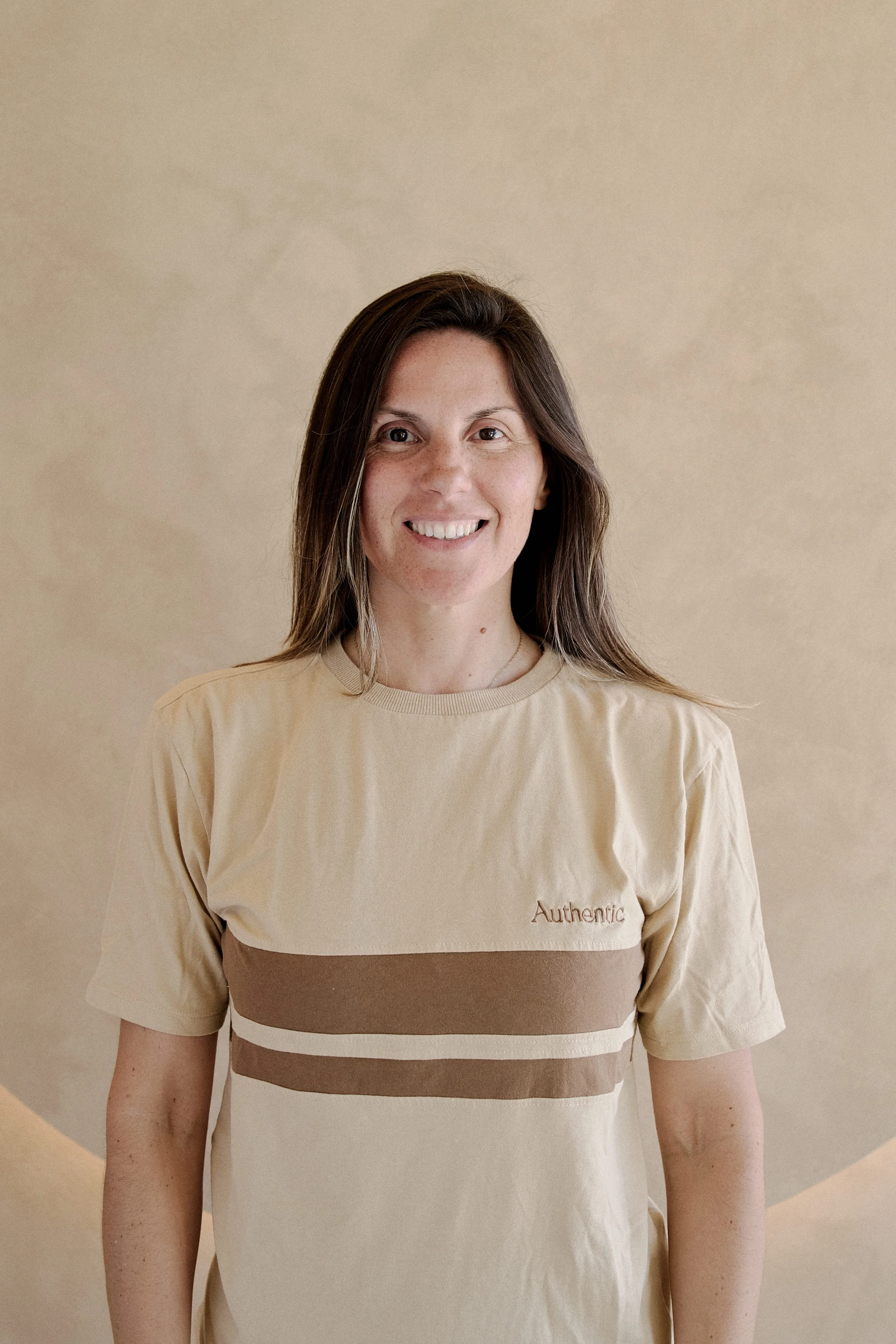 Mulher instrutora de Yoga do Authentic Premium Studio , usando uma camiseta bege com faixa castanha e escrito "Authentic" no lado esquerdo do peito, em pé contra um fundo bege.