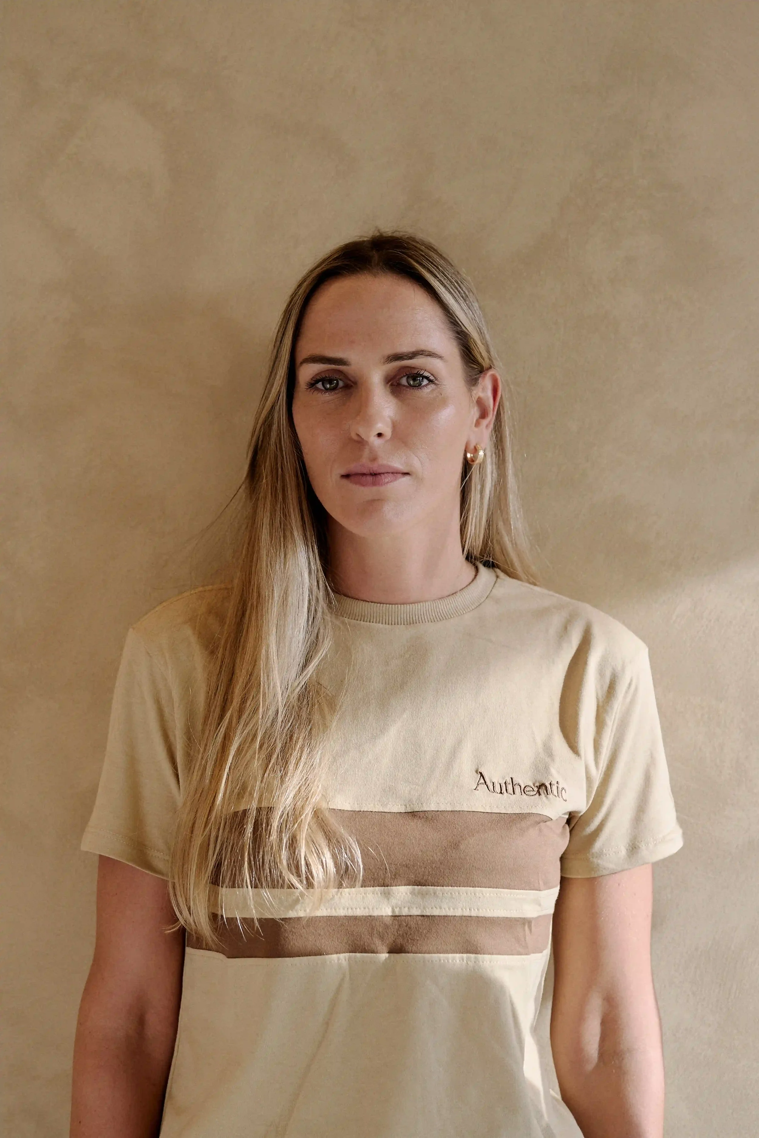 Mulher Nutricionista do Authentic Premium Studio , usando uma camiseta bege com detalhes em marrom, posando contra uma parede bege.