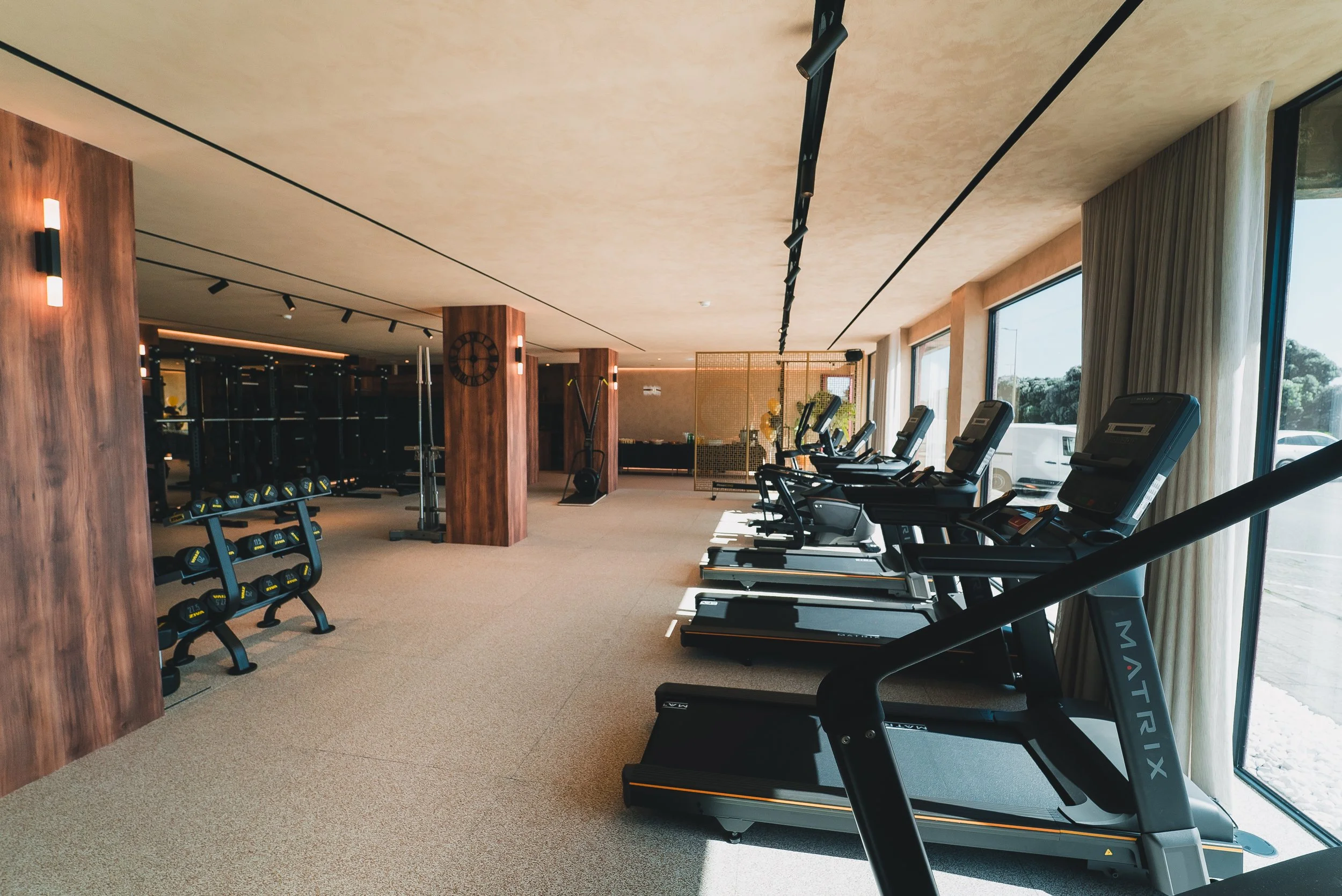 Sala de ginástica com esteiras de lado à janela, equipamentos de musculação ao fundo e parede de madeira.