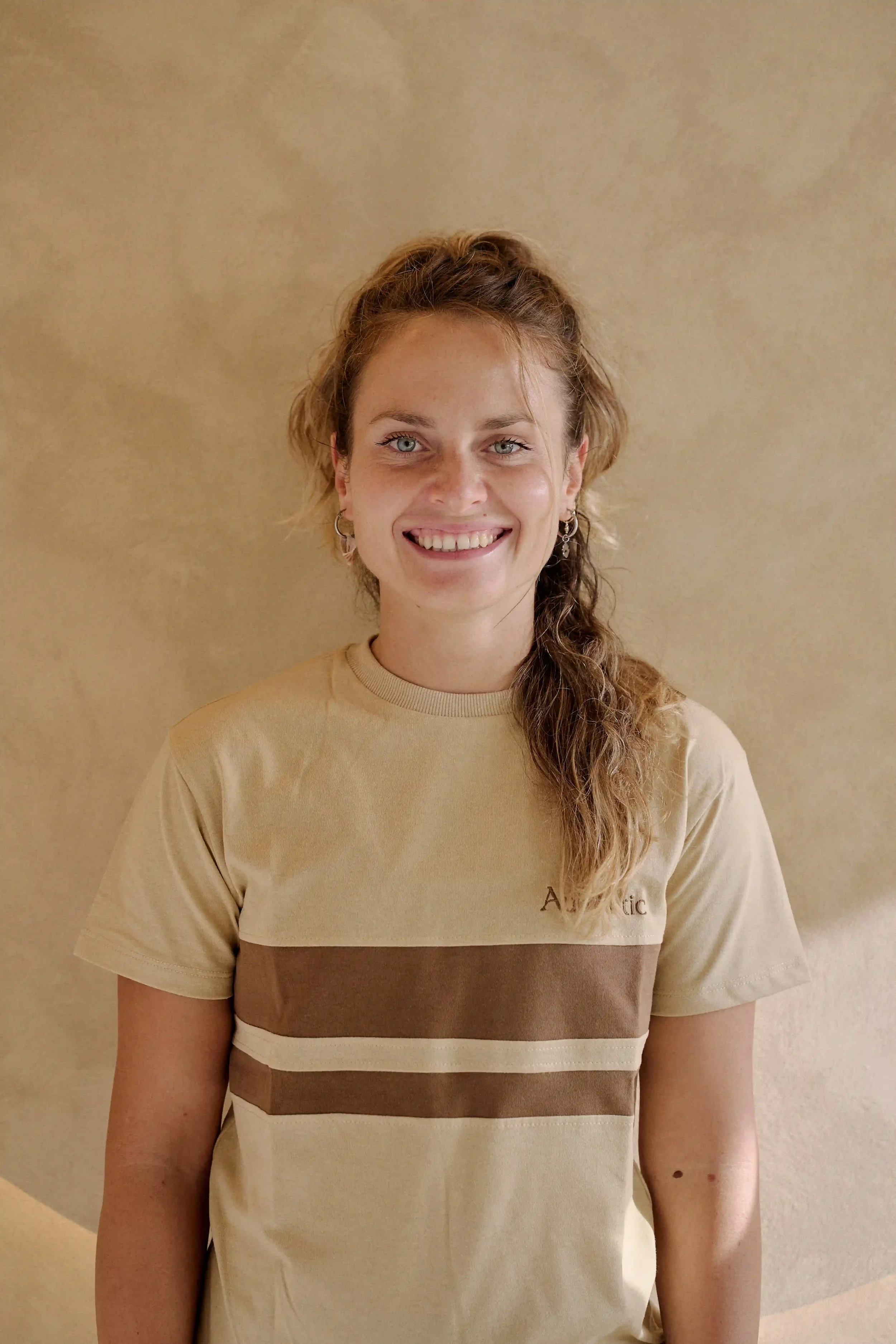 Mulher Professora de Barre e Yoga e Pilates do Authentic Premium Studio, usando camiseta bege com faixas marrons e creme, de brincos pequenos, posando contra parede bege.