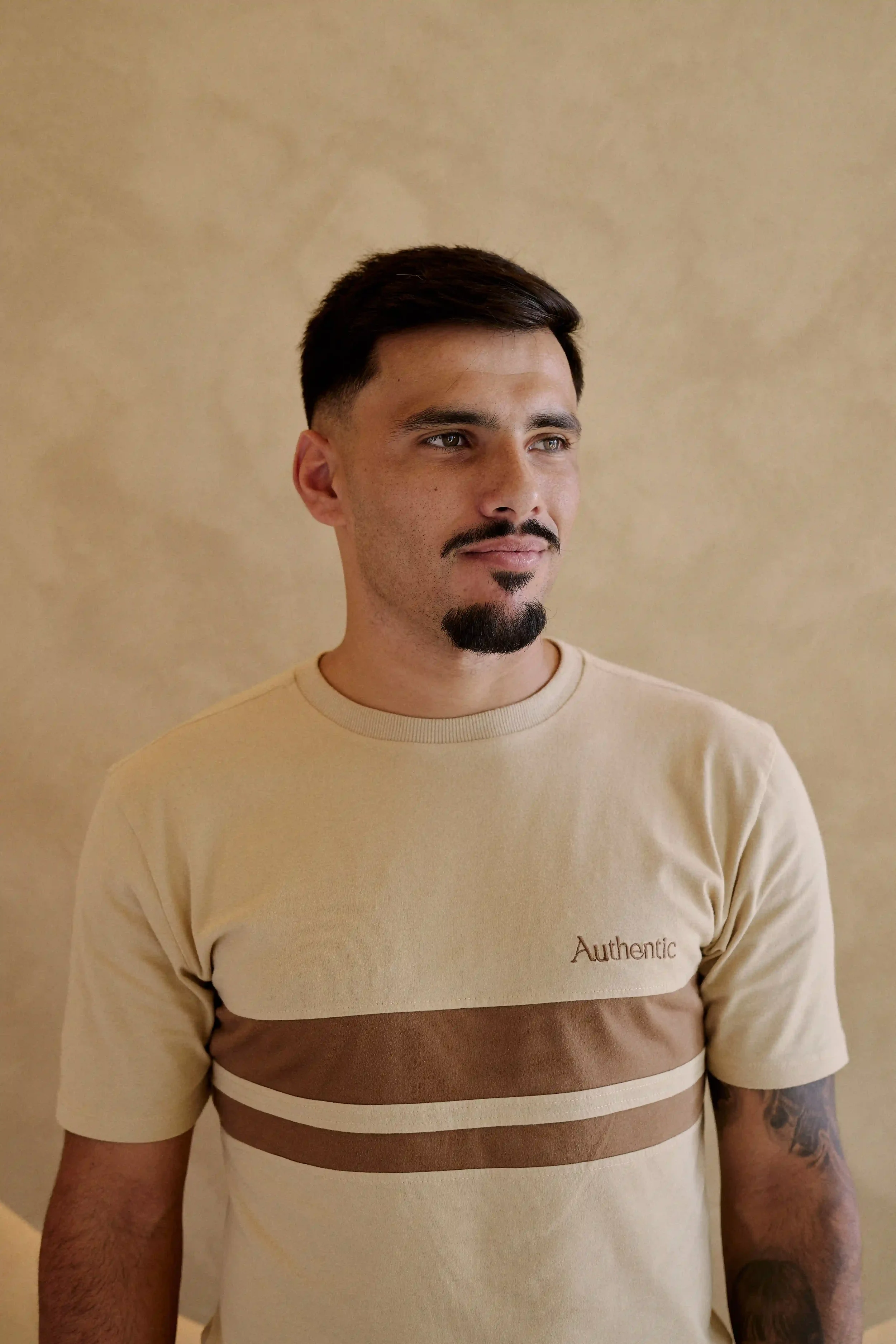 Homem Personal Trainer do Authentic Premium Studio, vestindo camiseta bege com faixas marrons, olhando para a câmera, parado contra uma parede bege. 