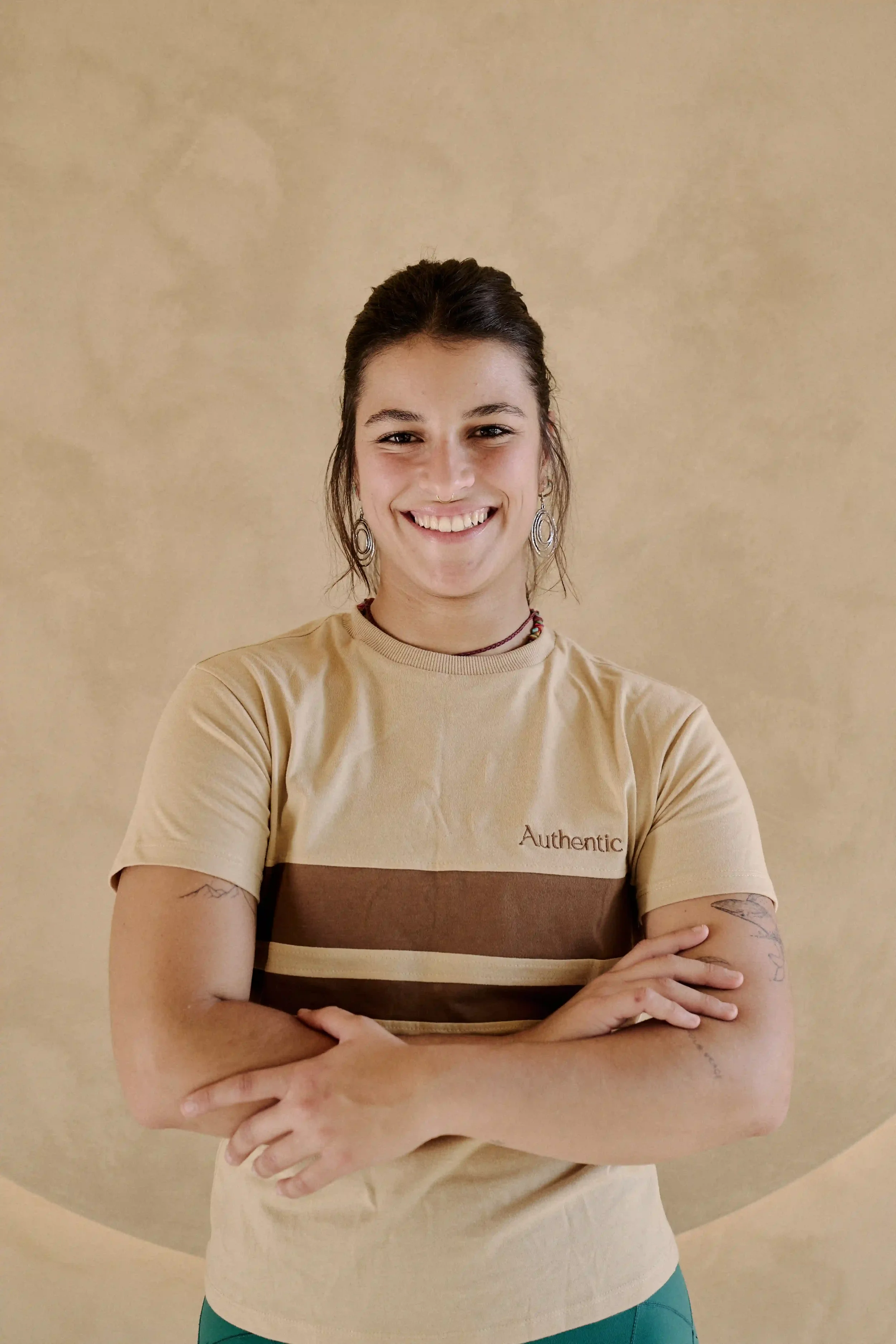 Mulher a sorrir com camiseta beje com a palavra "Authentic" escrito, parada em frente a parede bege