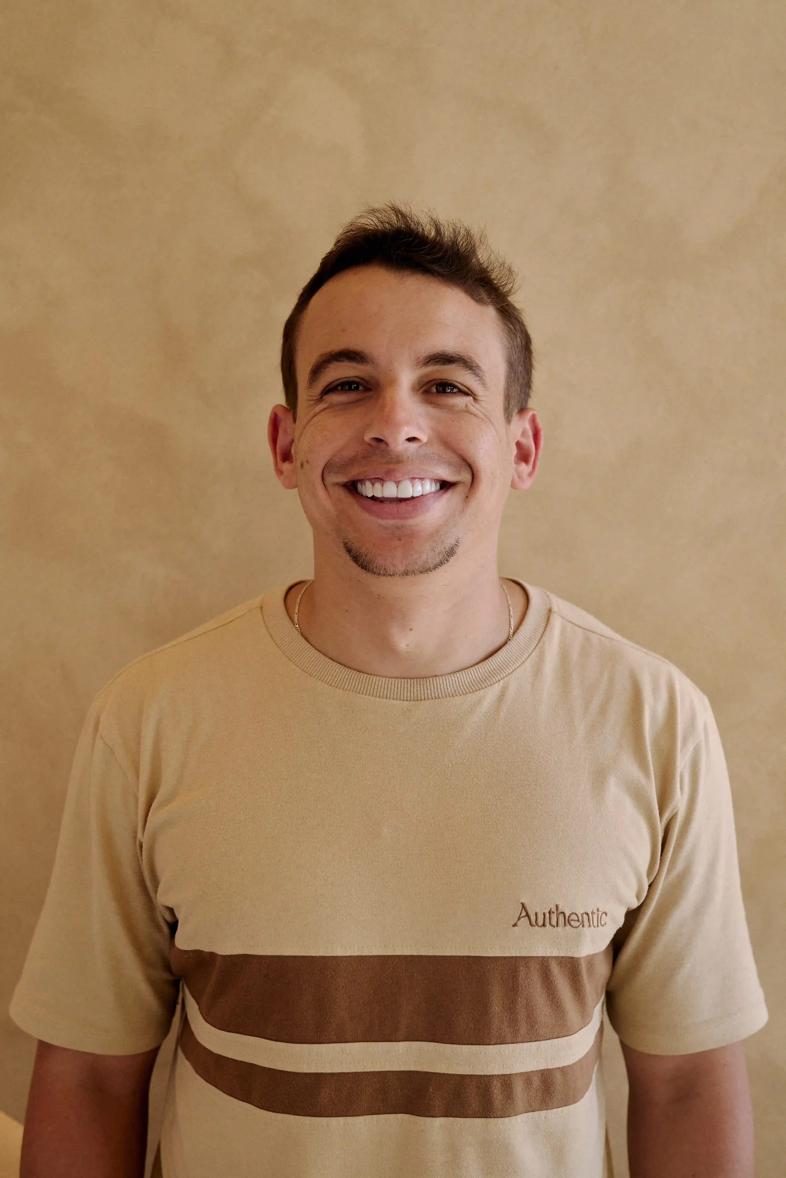 Homem Personal Trainer do Authentic Premium Studio, vestindo camiseta bege com faixas marrons, olhando para a câmera, parado contra uma parede bege.