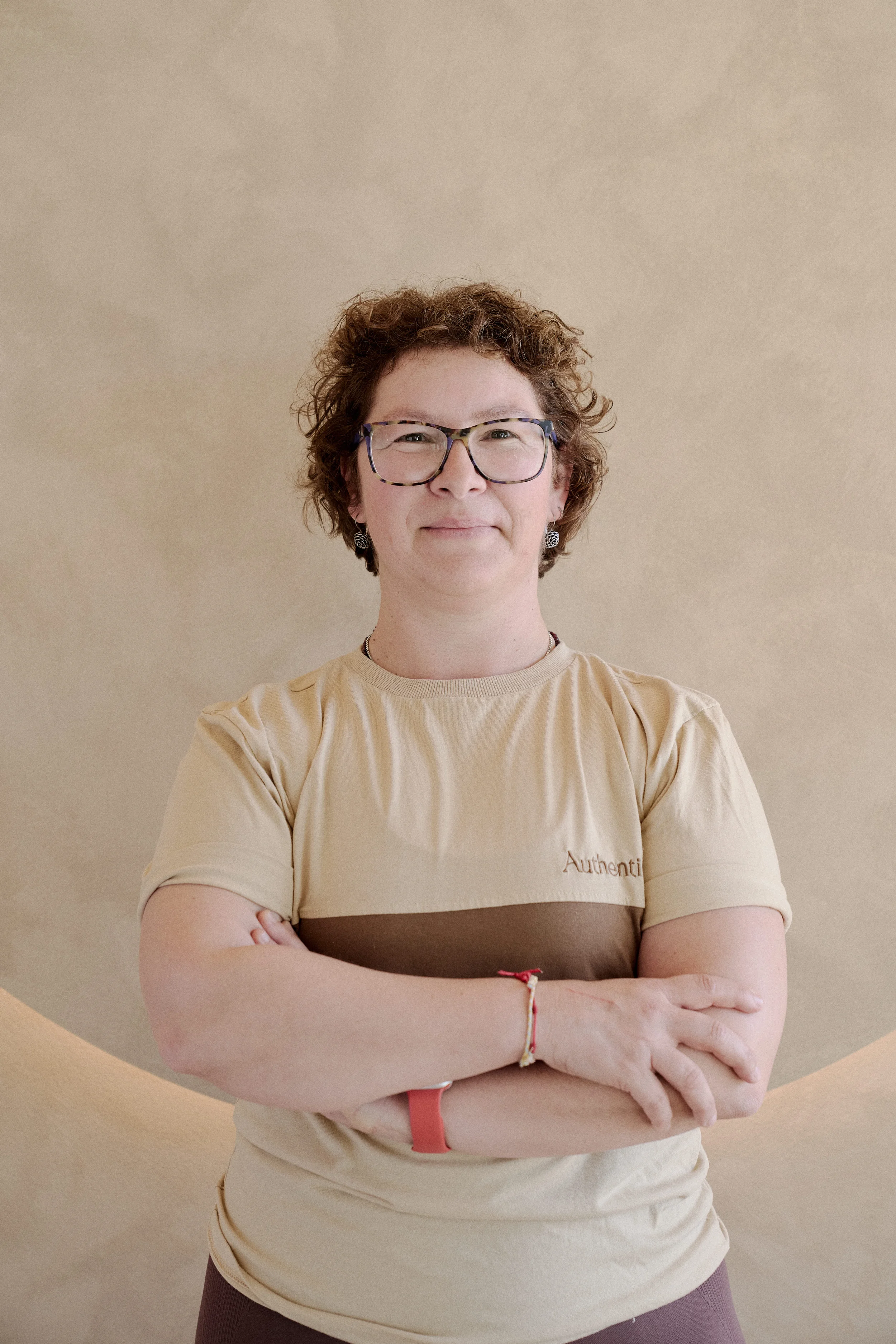 Mulher Instrutora de Pilates do Authentic Premium Studio , usando uma camiseta bege com faixa marrom e escrito "Authentic" no lado esquerdo do peito, em pé contra um fundo bege.