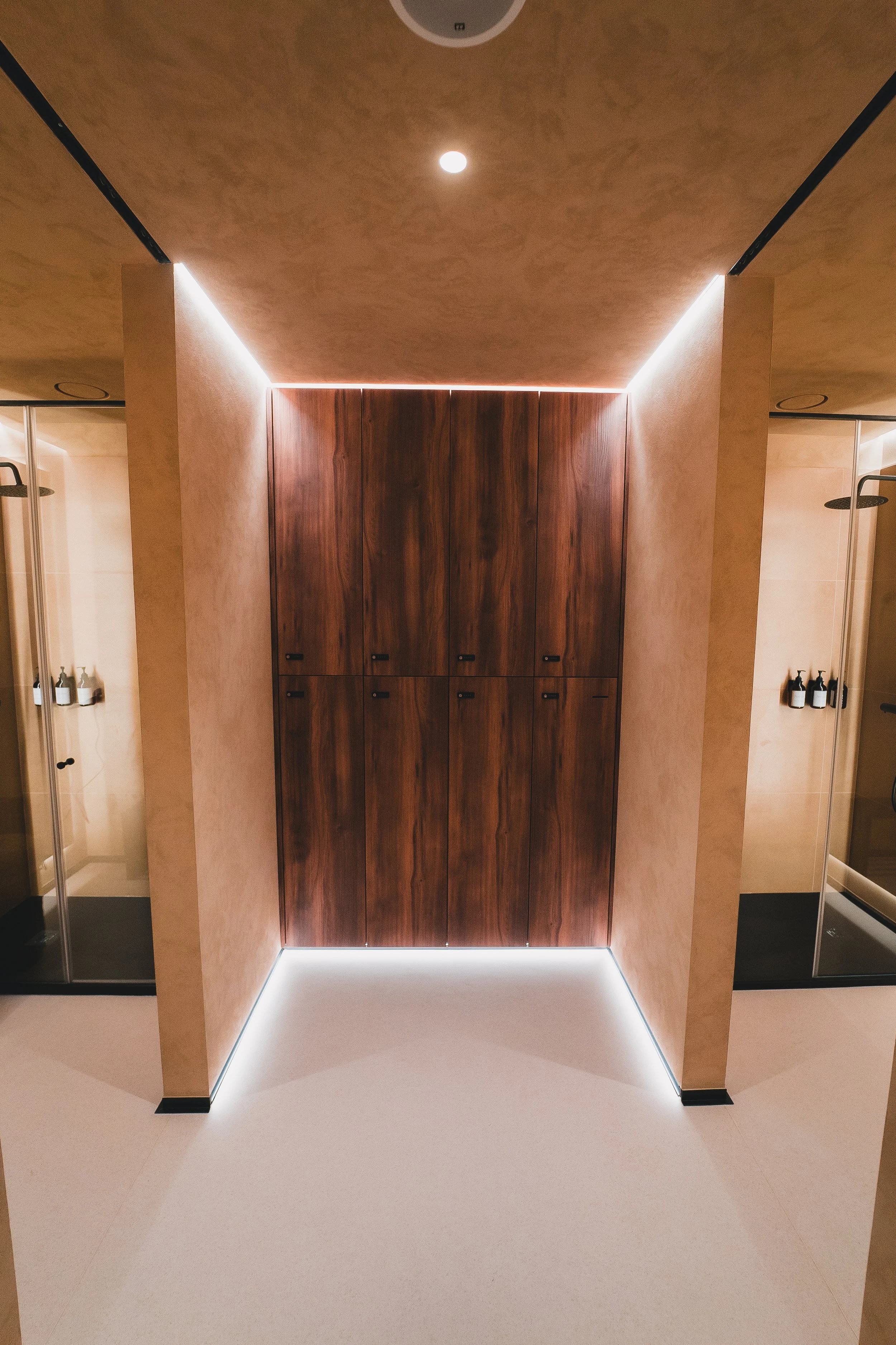 Imagem de um corredor com paredes de cor bege e iluminação LED discreta, que leva a um armário de madeira escura ao fundo e duas cabines de banho ou vestiários com portas de vidro ao lado.