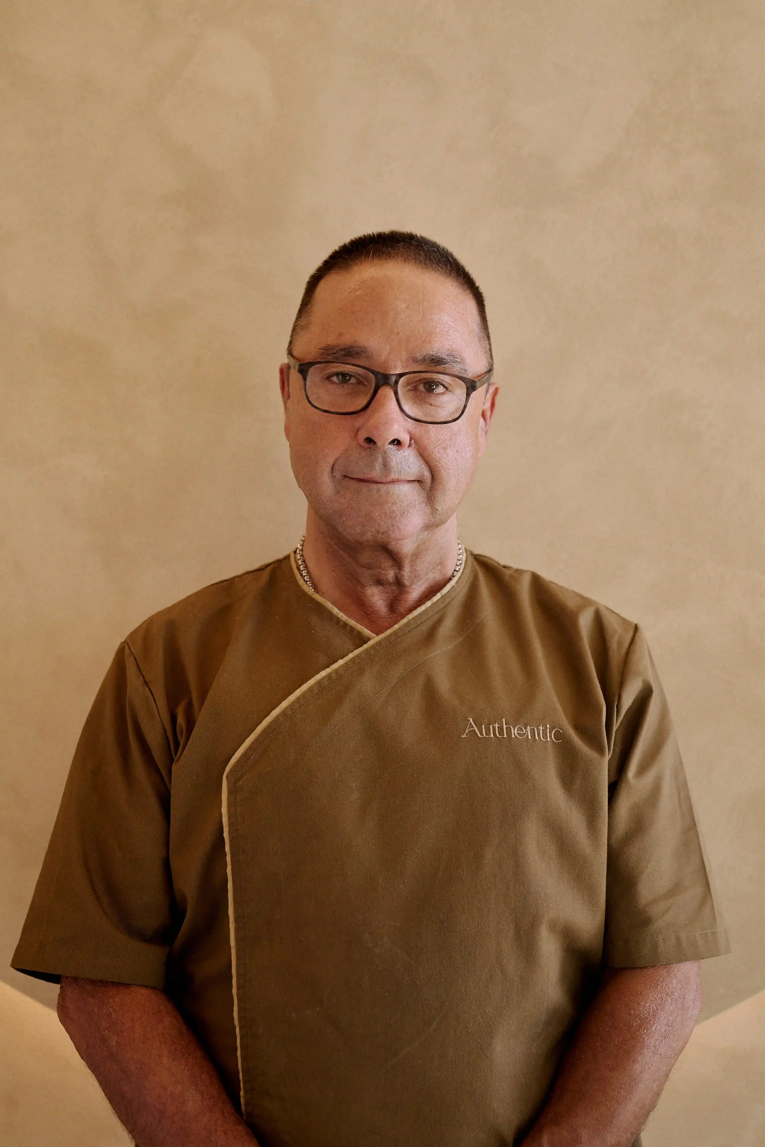 Homem Massoterapeuta, especialista em massoterapia e recuperação física do Authentic Premium Studio, usando uniforme marrom com a palavra 'Authentic' bordada, posando contra parede bege.