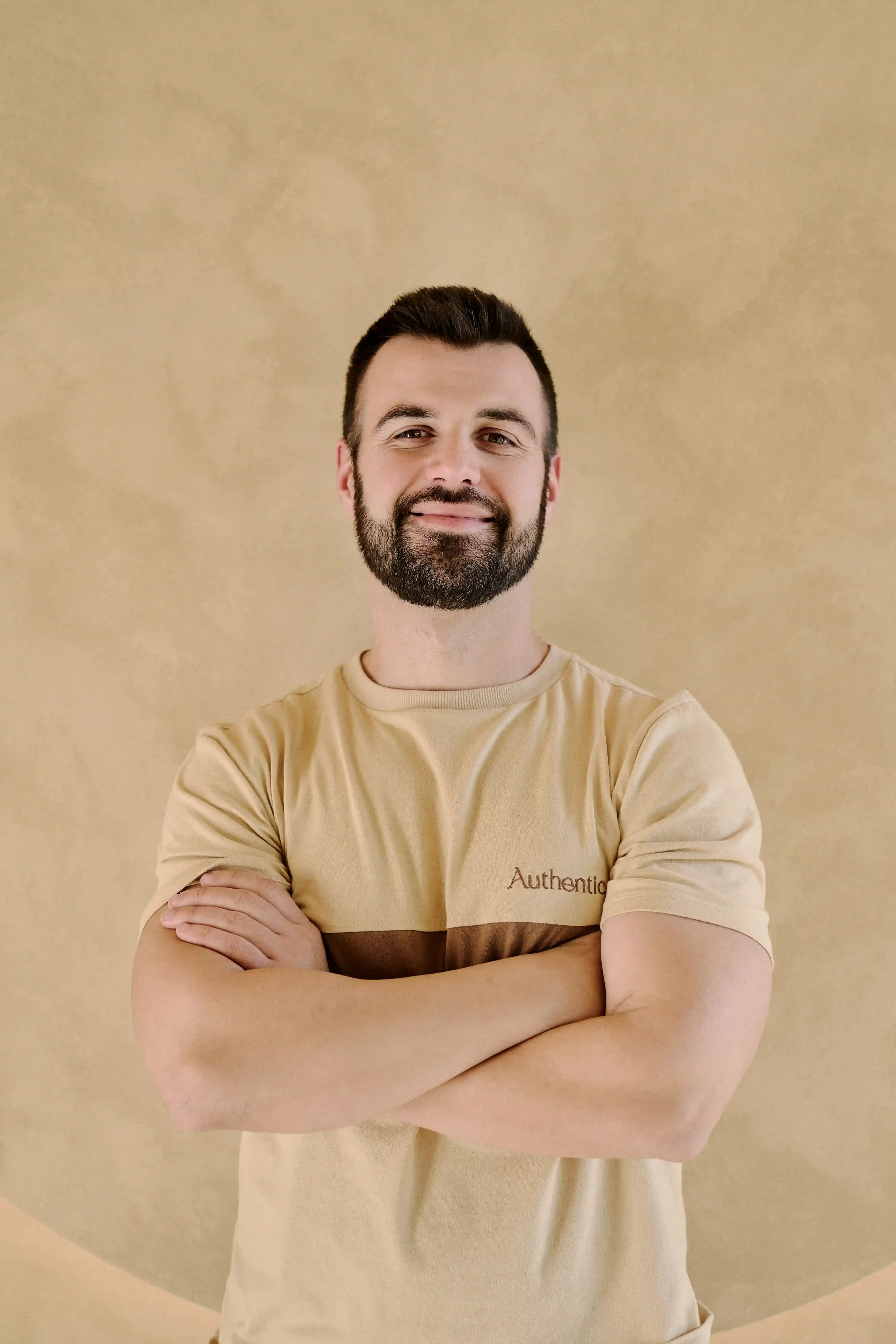 Homem Personal Trainer do Authentic Premium Studio com barba sorrindo, usando camiseta bege com faixas marrons e a palavra 'Authentic' escrita.