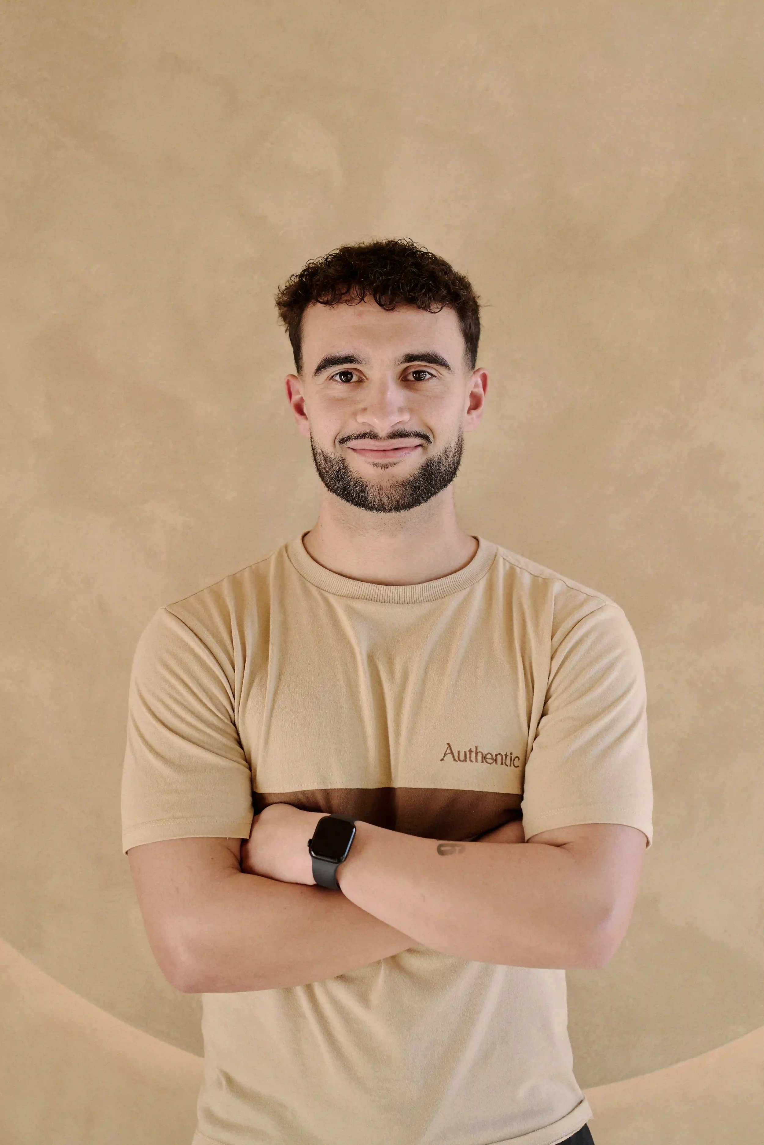 Homem Personal Trainer do Authentic Premium Studio, vestindo camiseta bege com faixas marrons, olhando para a câmera, parado contra uma parede bege. 